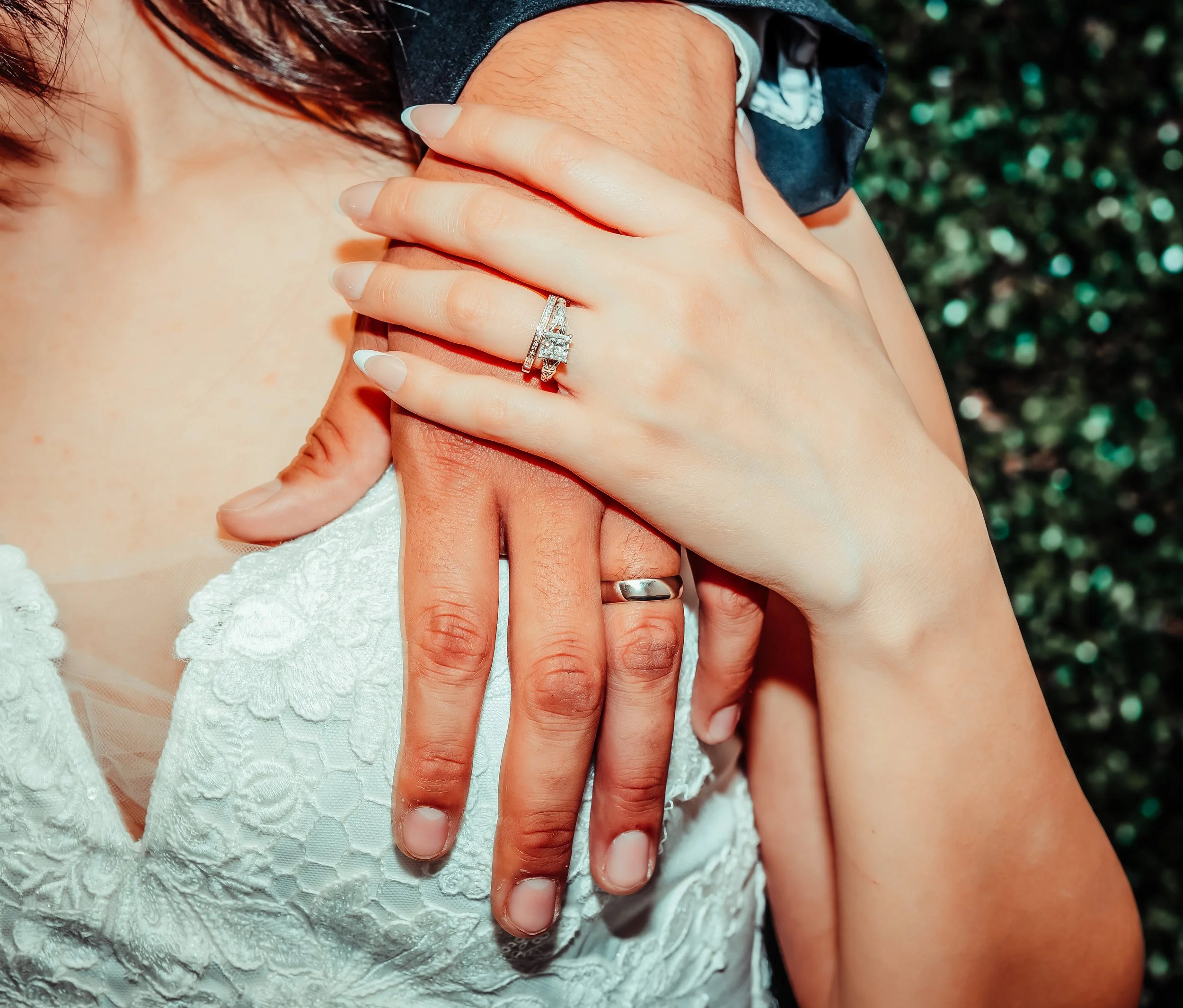 Close-up of two hands, one adorned with wedding rings, resting on a woman’s shoulder. The woman is wearing a white lace dress, and the background appears to be greenery. indoor Mexican wedding photographer phoenix az