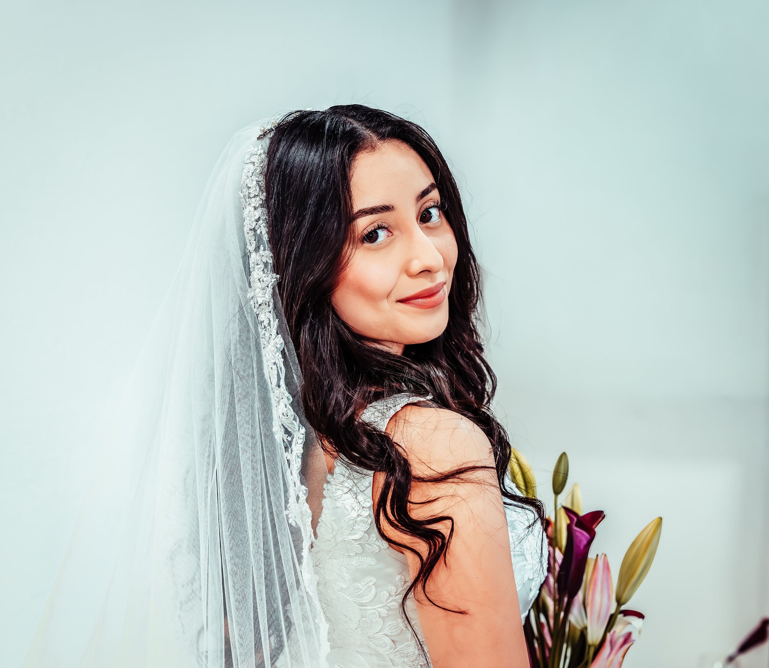 Young woman with long dark hair in a bridal gown and veil, holding pink lilies, smiling softly against a plain background. indoor Mexican wedding photographer phoenix az