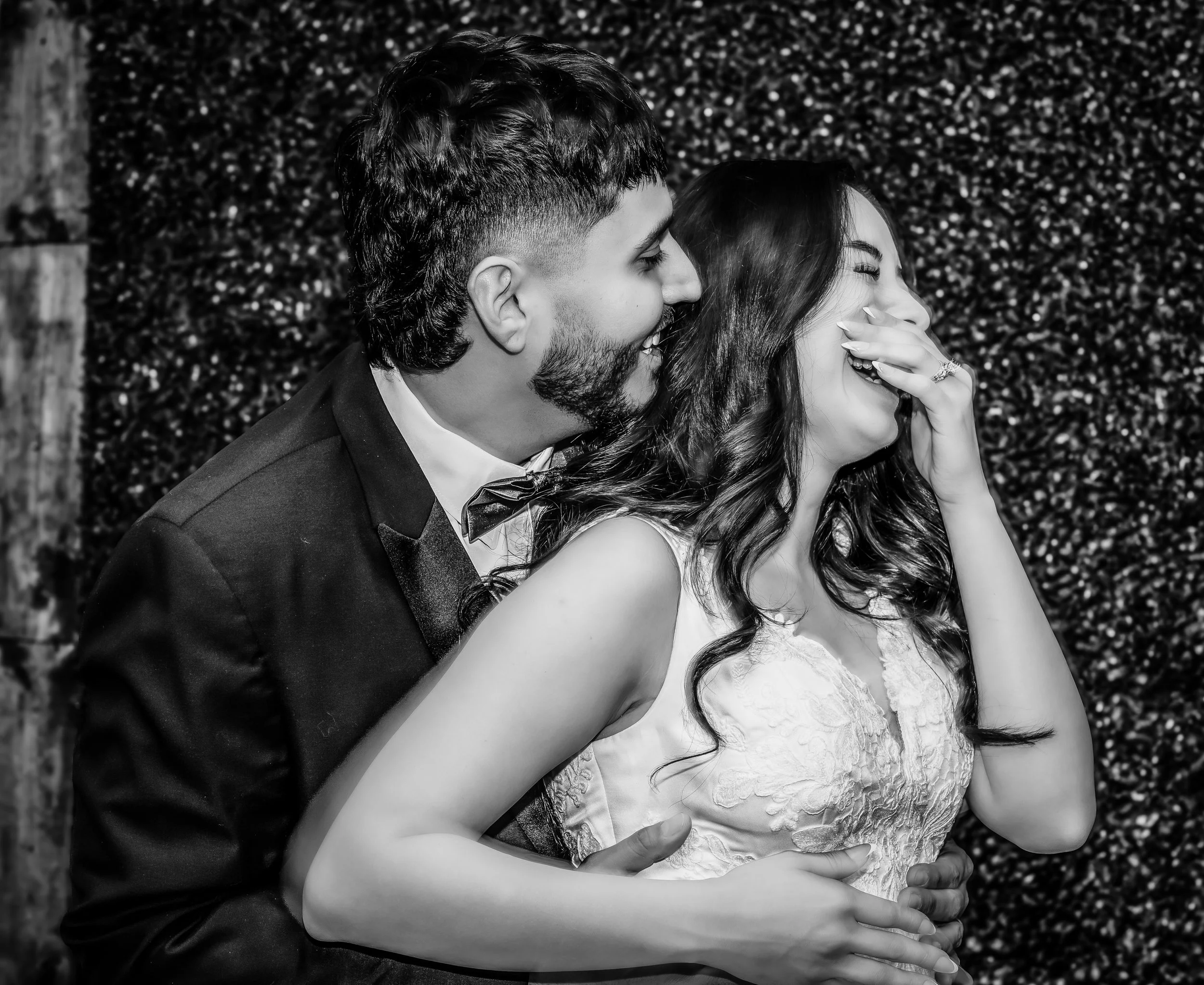 A black-and-white photo of a happy couple, possibly at a wedding, sharing a joyful moment with the man in a tuxedo and the woman in a lace dress, both laughing and embracing. indoor Mexican wedding photographer phoenix az
