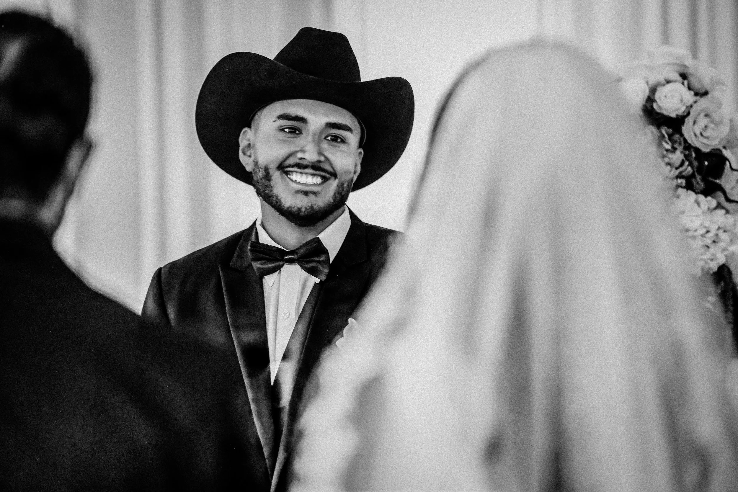 A man wearing a cowboy hat, tuxedo, and bow tie smiling during a wedding ceremony. The bride, with a veil and floral headpiece, and another person are partially visible in the foreground. indoor Mexican wedding photographer phoenix az