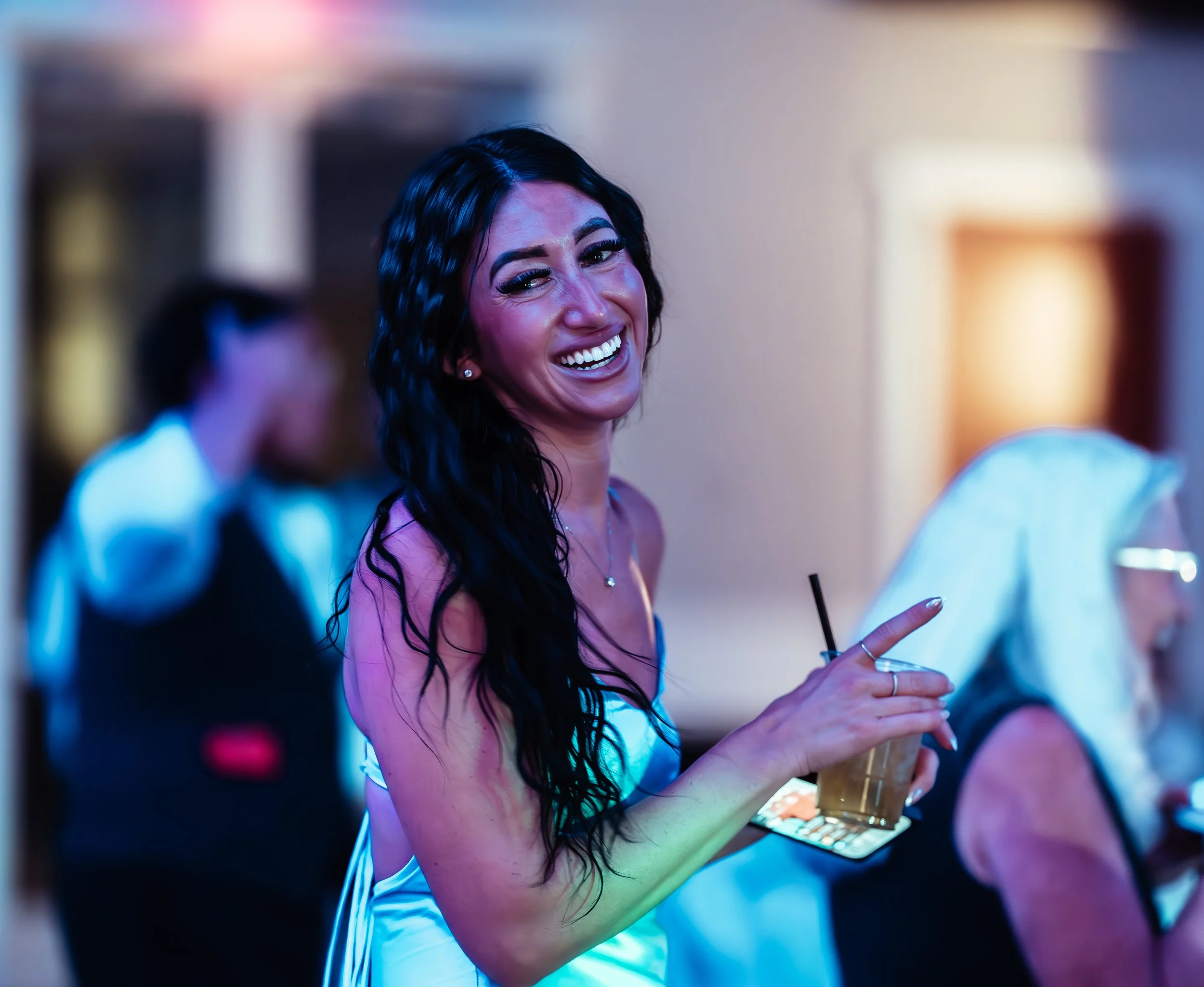 A woman with long black curly hair smiling and holding a drink at a social event, with other people blurred in the background. western wedding photographer az rustic wedding photographer az barn wedding photographer az backyard wedding photographer a