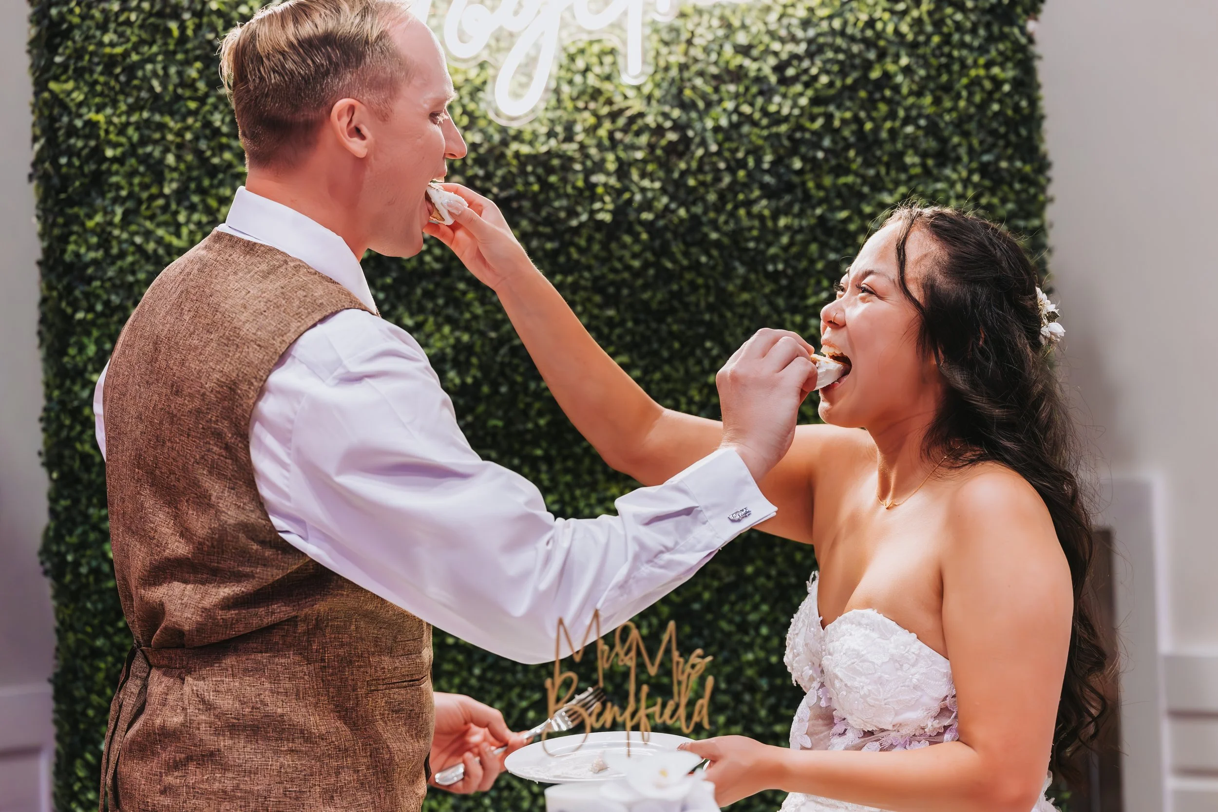 Bride and groom sharing cake during wedding celebration, with green hedge backdrop and 'Mr & Mrs' cake topper. western wedding photographer az rustic wedding photographer az barn wedding photographer az backyard wedding photographer az