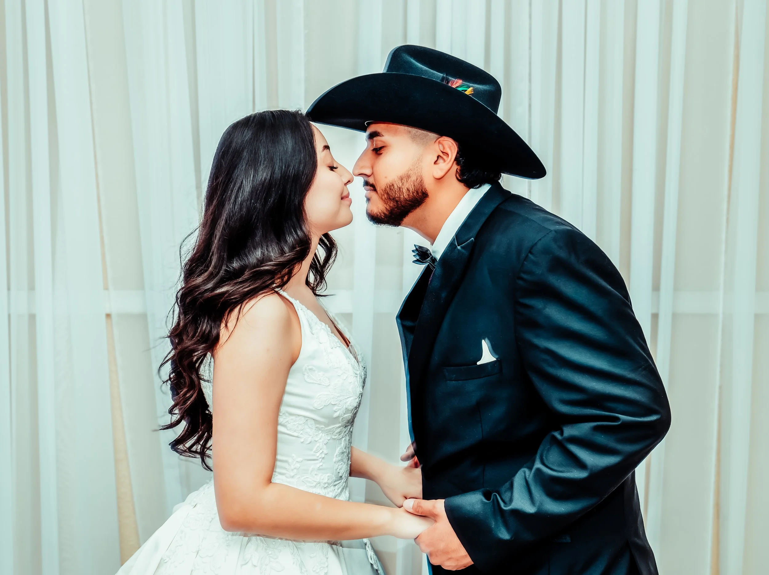 A newlywed couple, a woman in a white wedding dress and a man in a black tuxedo and cowboy hat, holding hands and about to kiss, standing in front of cream-colored curtains. indoor Mexican wedding photographer phoenix az