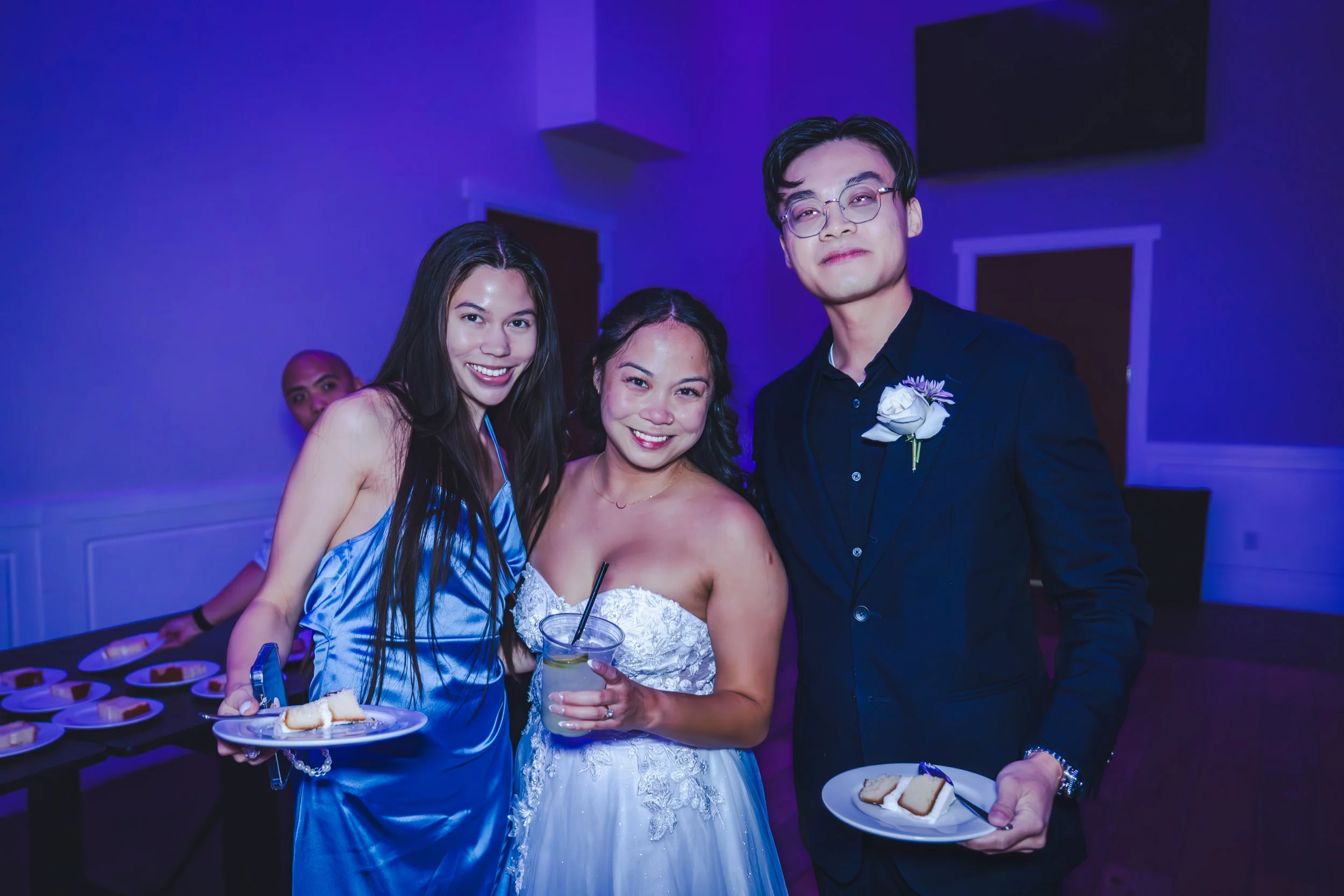 Group of three young adults at a wedding reception, standing together with smiles, dressed in formal attire, holding plates of cake and drinks, with a person in the background and a purple lit room. western wedding photographer az rustic wedding phot