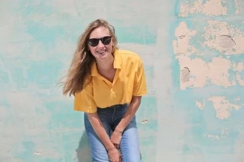 smiling blond young woman wearing yellow shirt leaning against a wall
