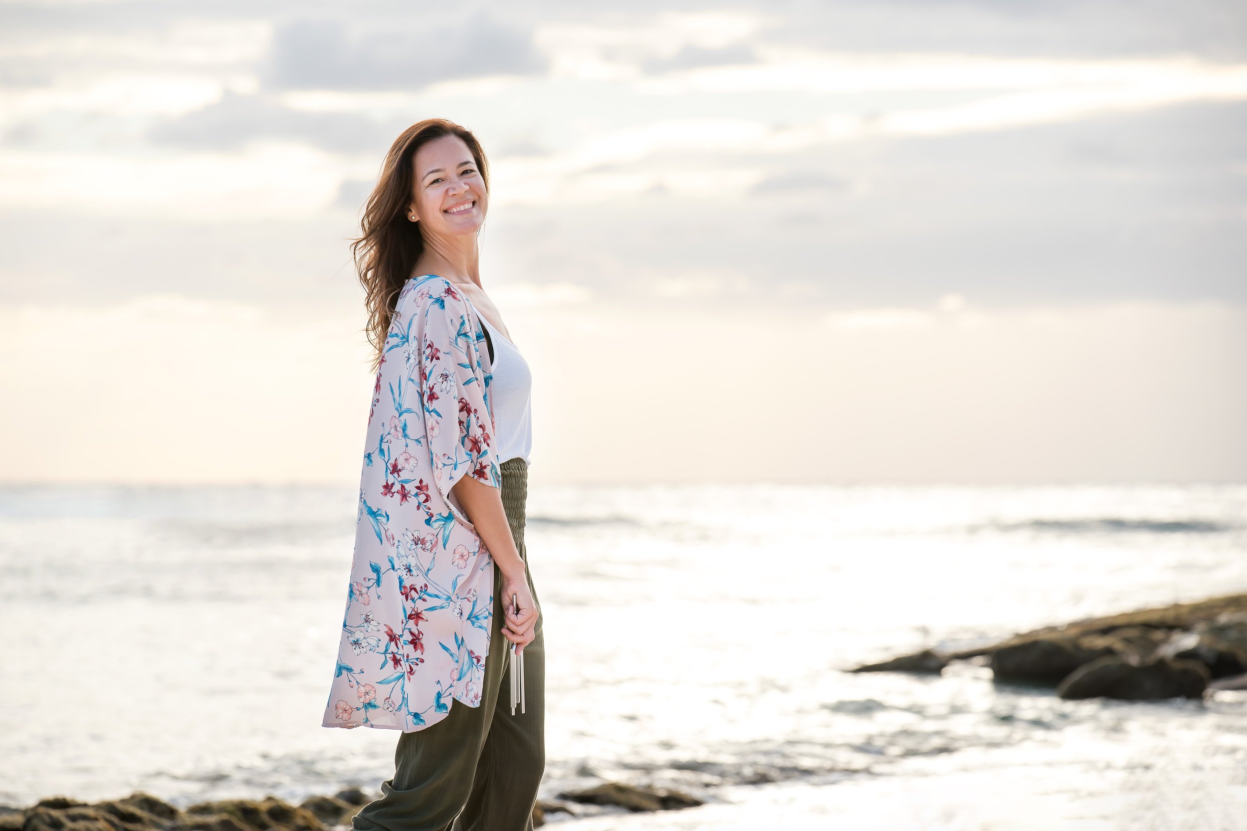 A woman standing on a rocky shoreline with the ocean and cloudy sky in the background, smiling at the camera.