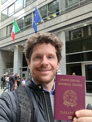 Hombre sosteniendo un pasaporte europeo frente a un edificio con banderas de Italia y la Unión Europea.