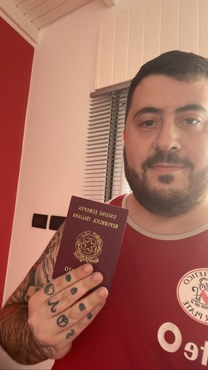 Un hombre sosteniendo un pasaporte italiano frente a una ventana con persianas. Lleva una camiseta sin mangas roja y tiene tatuajes en el brazo izquierdo.