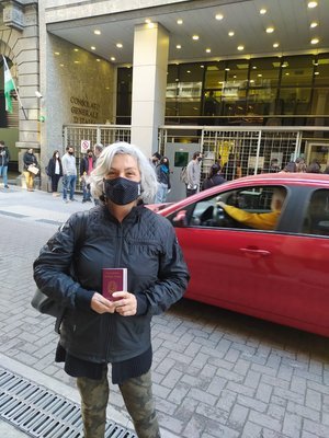 Persona frente al edificio "Consejo Profesional de Ciencias Económicas", sosteniendo un documento en la mano y llevando una mascarilla. Un auto rojo pasa por la calle.