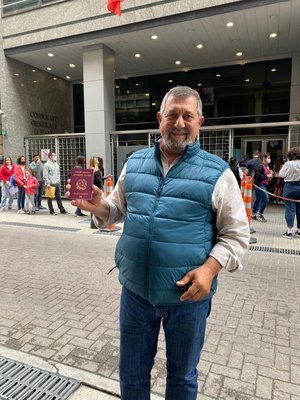 Hombre sostiene un pasaporte frente a un edificio con fila de personas.