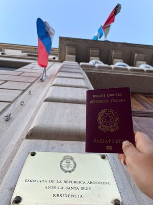 Persona sosteniendo un pasaporte italiano afuera de la Embajada de la República Argentina ante la Santa Sede, con banderas argentinas ondeando al fondo.