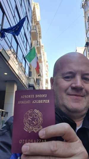 Hombre sosteniendo un pasaporte italiano frente a un edificio con banderas de la Unión Europea e Italia.