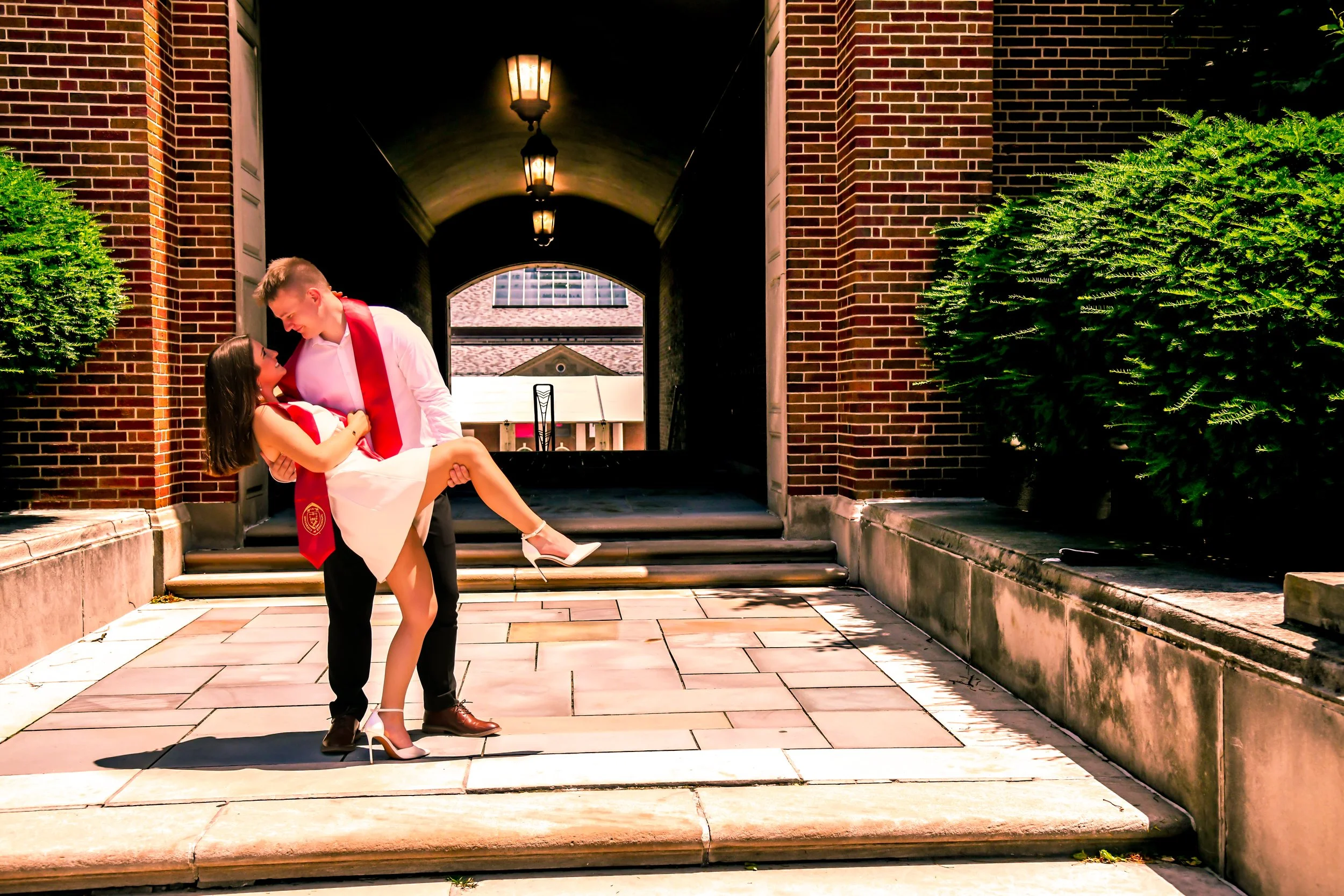 Couple dressed in graduation attire, with the woman being carried by the man, standing on a paved walkway outside a brick building with greenery.