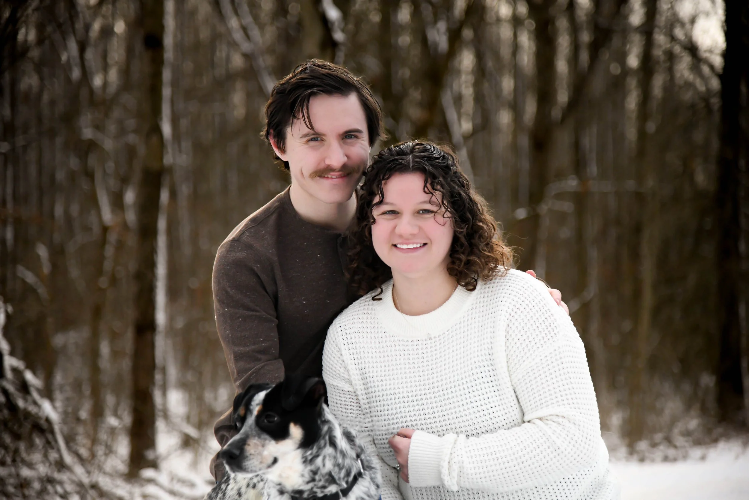 A smiling couple with a black and white dog in a snowy forest.