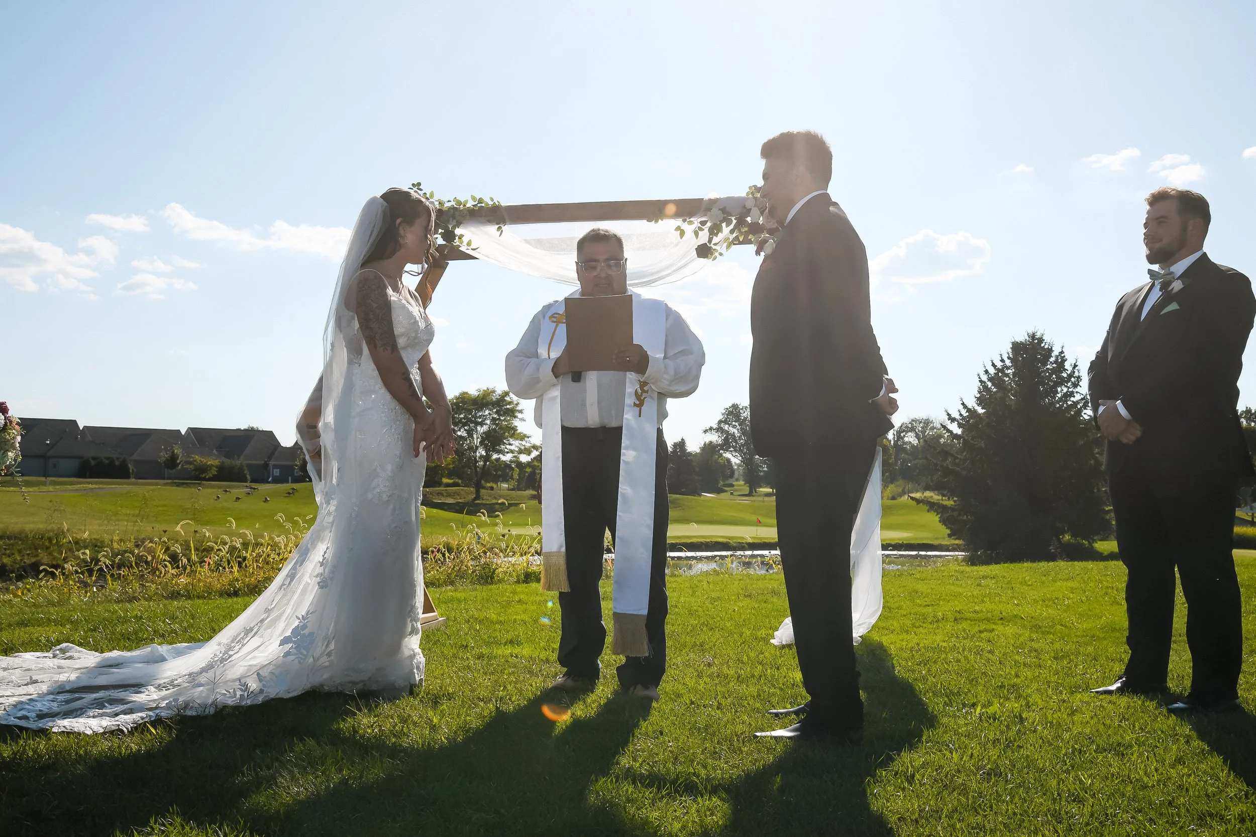 A couple getting married outdoors with a minister officiating, under a decorated arch, on a sunny day with green grass and trees in the background.