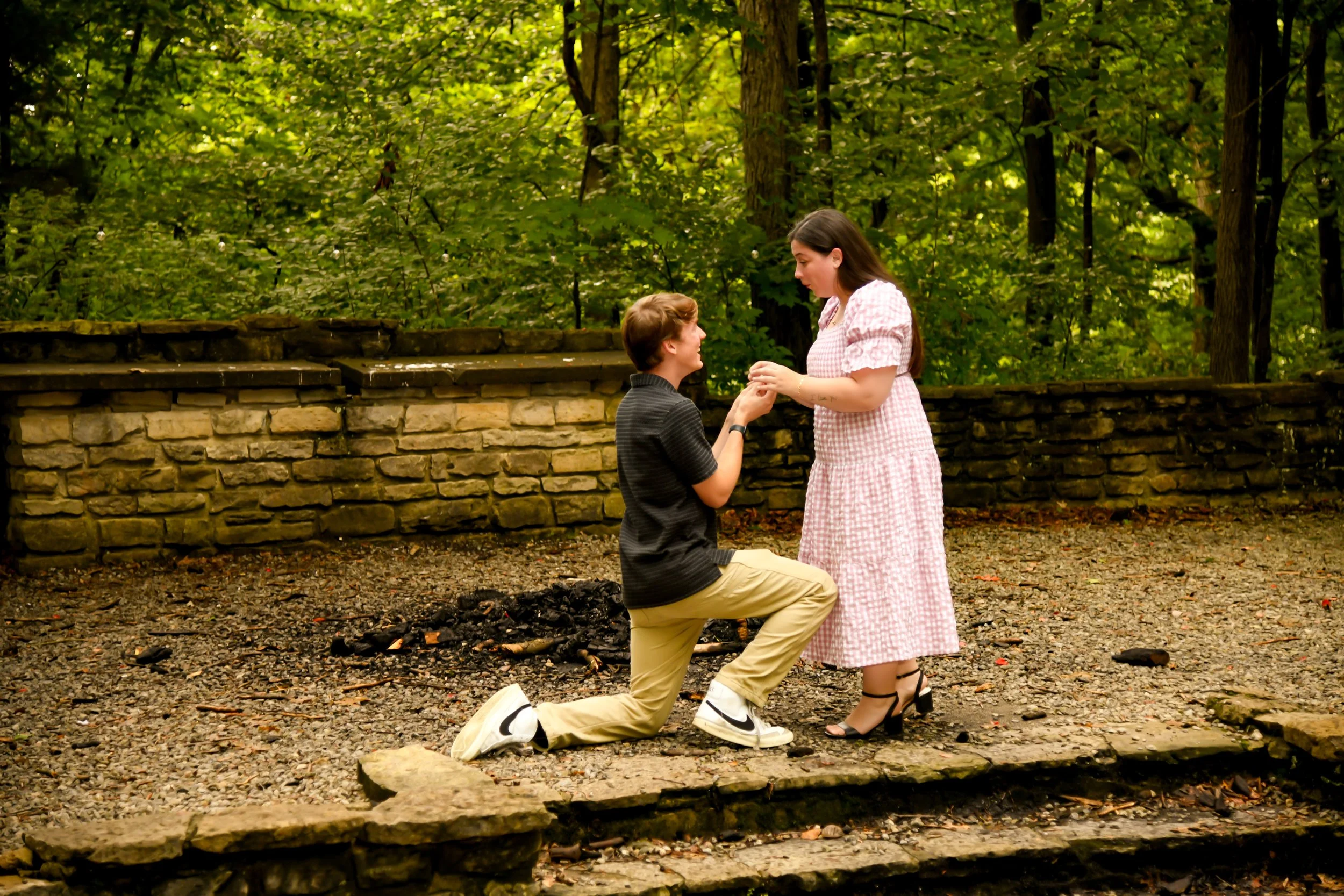 A man proposes marriage to a woman in a wooded outdoor setting, with a stone wall in the background. The man is kneeling on one knee, holding the woman's hands, while the woman looks down at him with surprise.