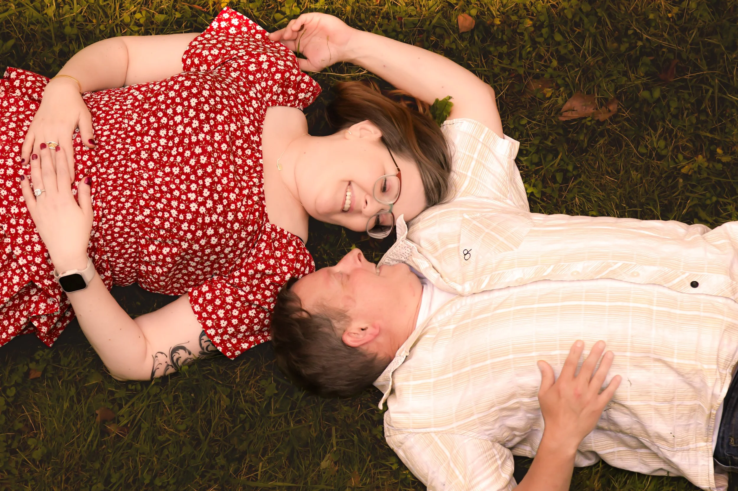 A woman and a man lying on the grass, smiling and looking at each other. The woman is wearing glasses and a red floral dress, with a gold bracelet and rings. The man is wearing a light-colored striped shirt and has a tattoo on his arm.