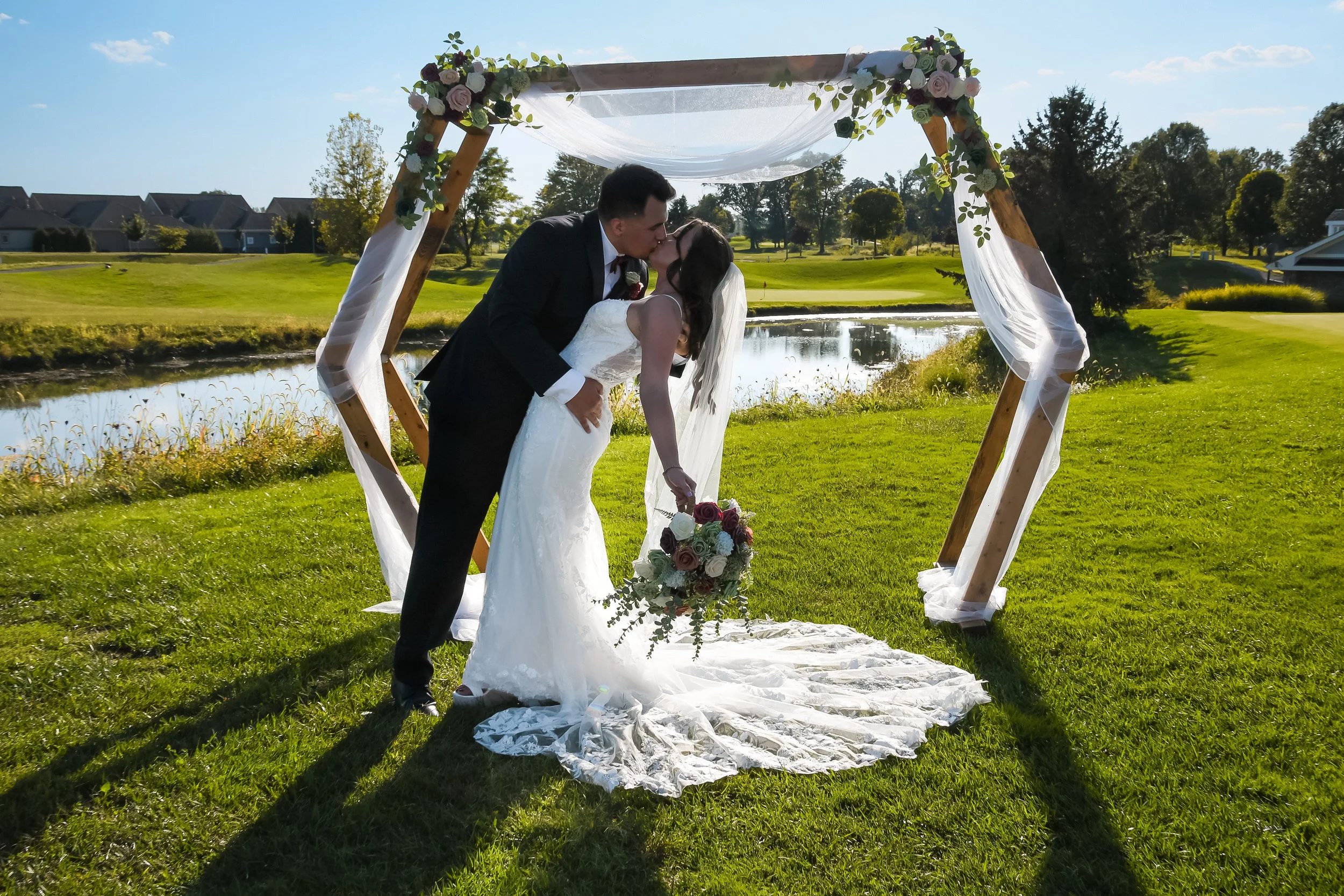 A newlywed couple sharing a kiss under a wooden wedding arch decorated with flowers and white fabric, outdoors near a pond on a sunny day.