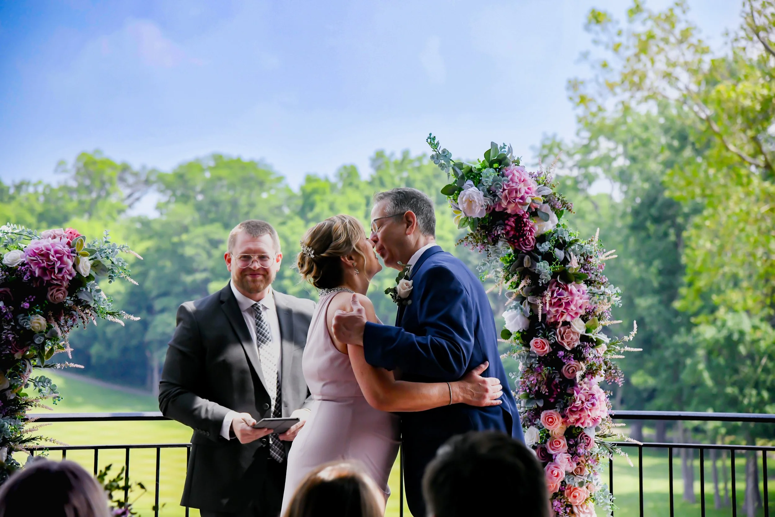 A wedding ceremony with a couple kissing under a floral arch, officiant standing nearby, outdoor setting with trees and greenery in the background.