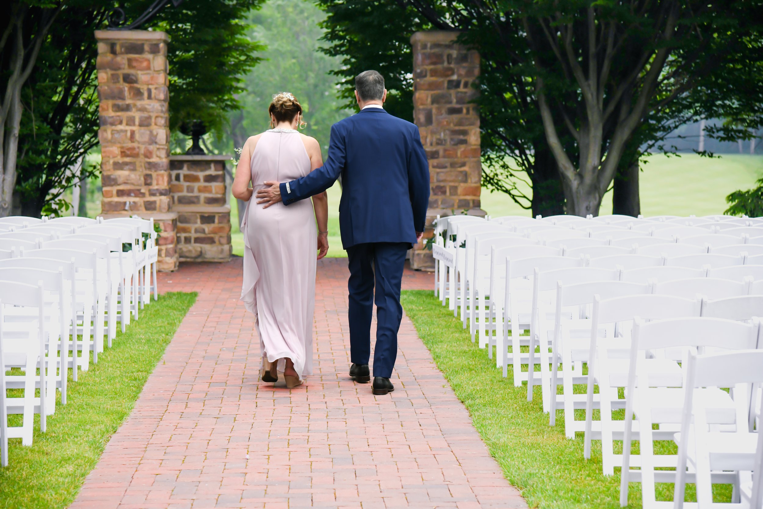 A couple walking down an outdoor aisle with white chairs on either side, surrounded by trees and brick columns, possibly at a wedding ceremony.