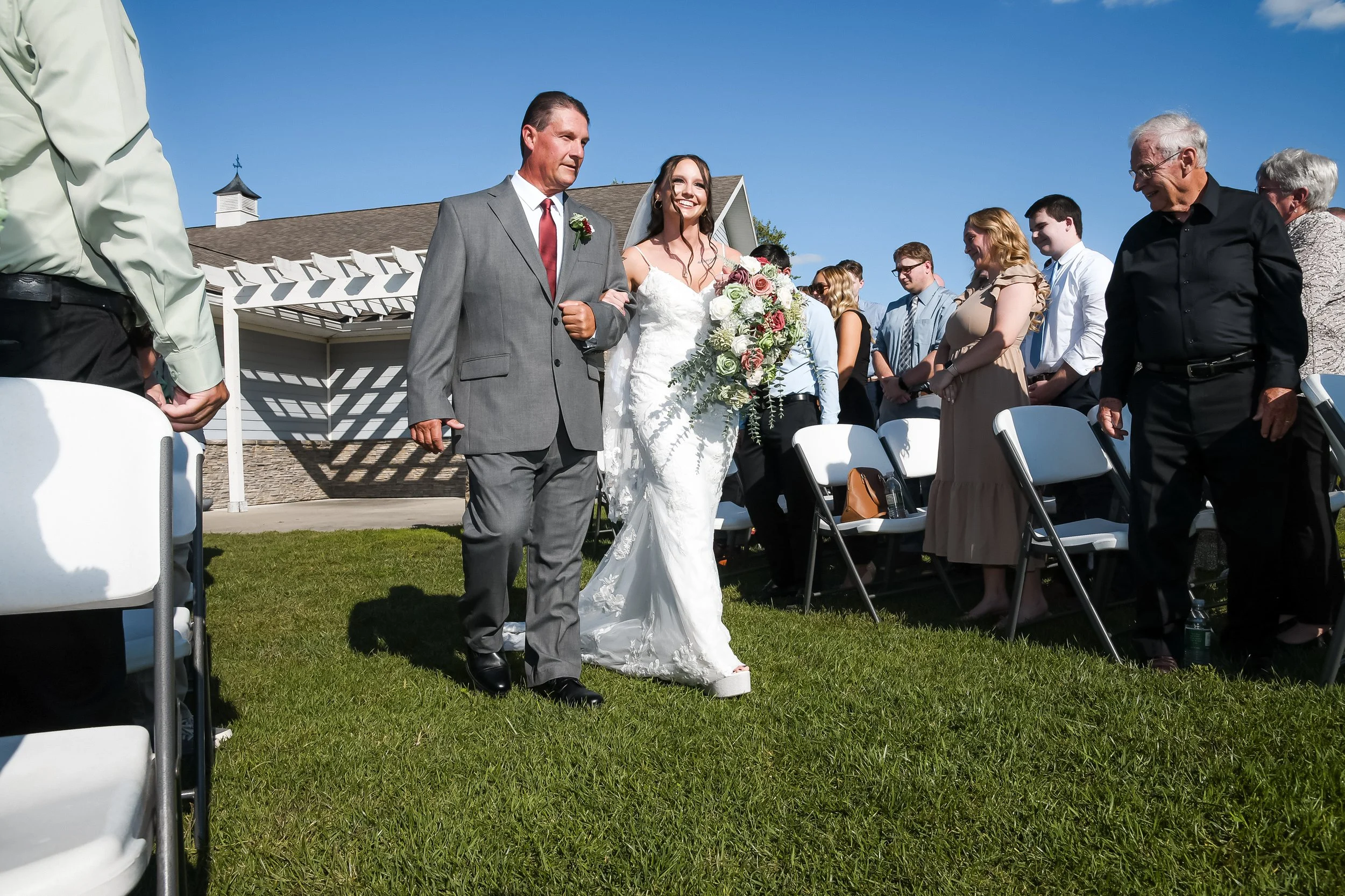 Bride in a white wedding dress walking down the aisle, accompanied by an older man in a gray suit, at an outdoor wedding ceremony on a sunny day.
