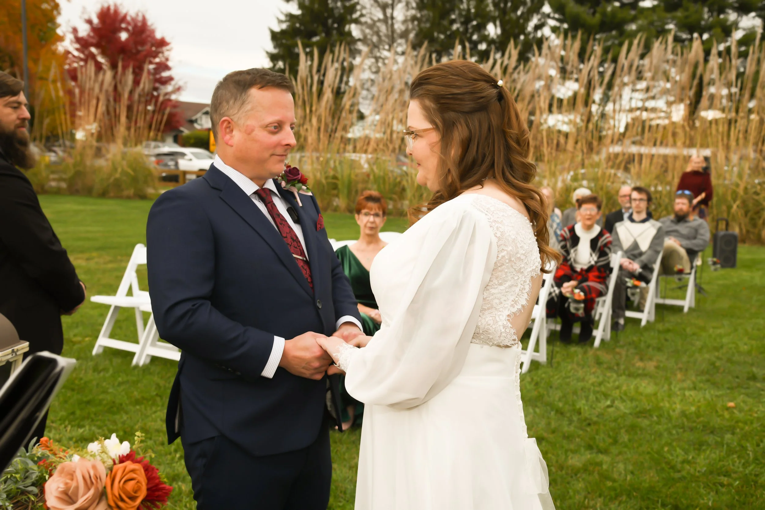 A wedding ceremony outdoors with a bride and groom holding hands and exchanging vows. The groom is wearing a navy suit and the bride is in a white wedding dress with lace details. Guests seated in the background and autumn trees are visible.
