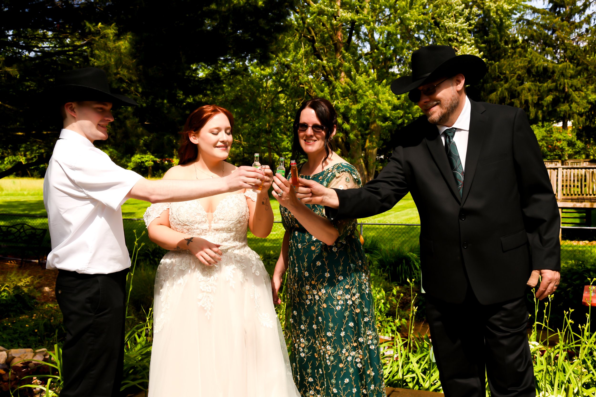 A group of four people, two men and two women, standing outdoors during the daytime. They are toasting with small bottles, smiling and enjoying the moment. The woman in a wedding dress is in the center, with the others dressed in semi-formal and casu