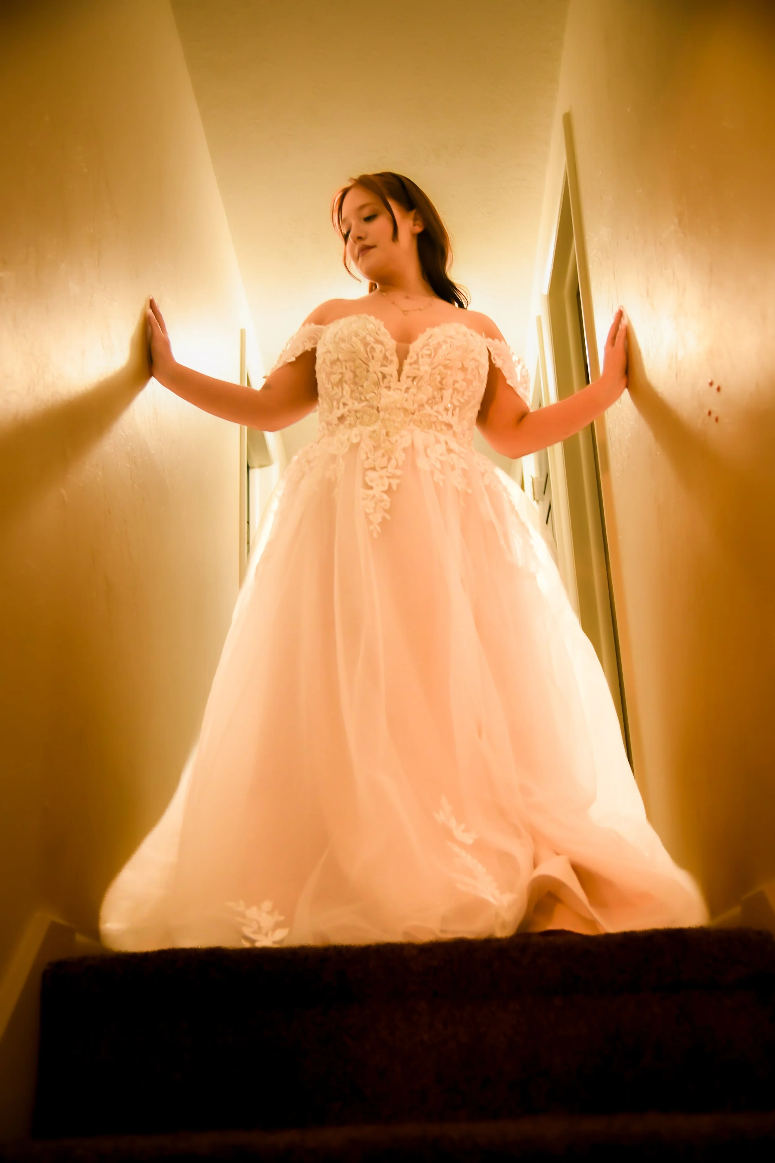 A woman in a light-colored, off-the-shoulder wedding dress with lace details is standing in a narrow hallway, looking down with her hands touching the walls.