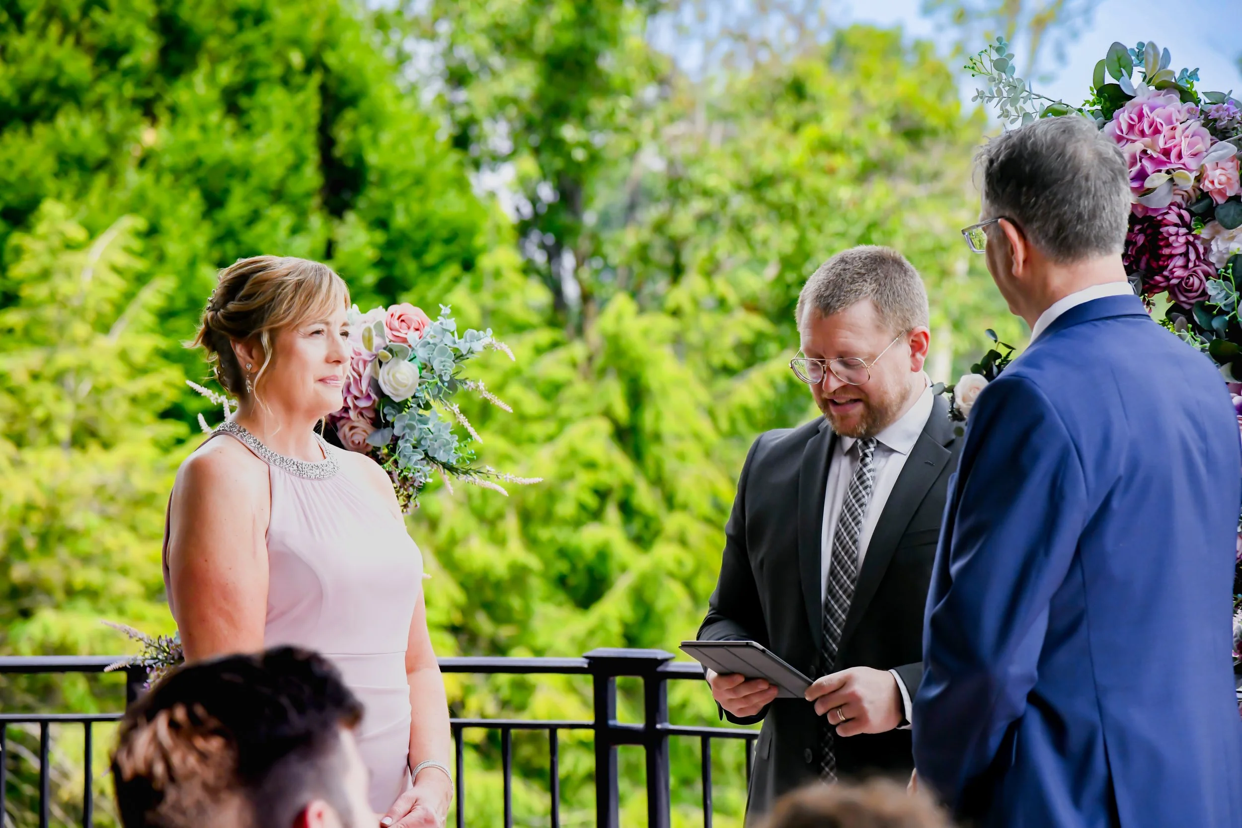 A woman in a light pink dress with embellishments on the neckline standing outdoors during a wedding ceremony, with a man in a dark suit and another man in a blue suit. The officiant is reading from a tablet, and the background features lush green tr