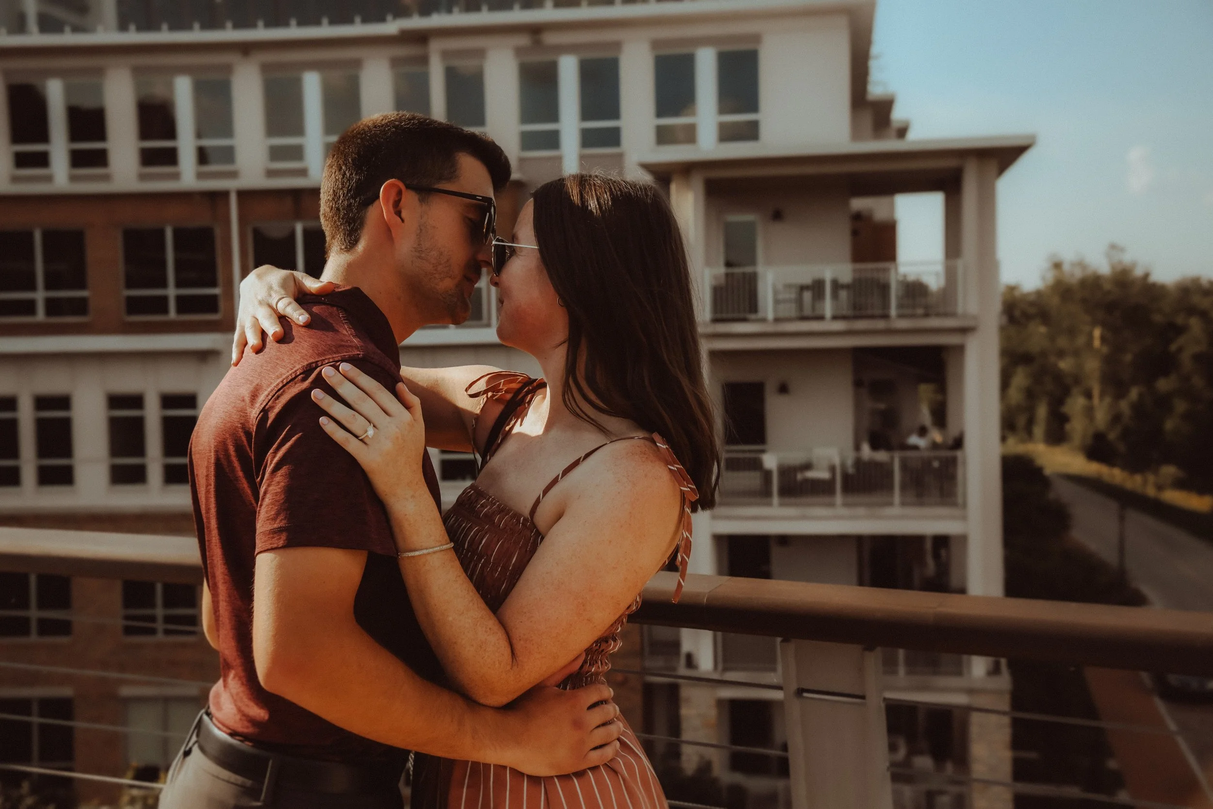 A couple is embracing and about to kiss on a balcony, with a modern apartment building in the background during sunset.