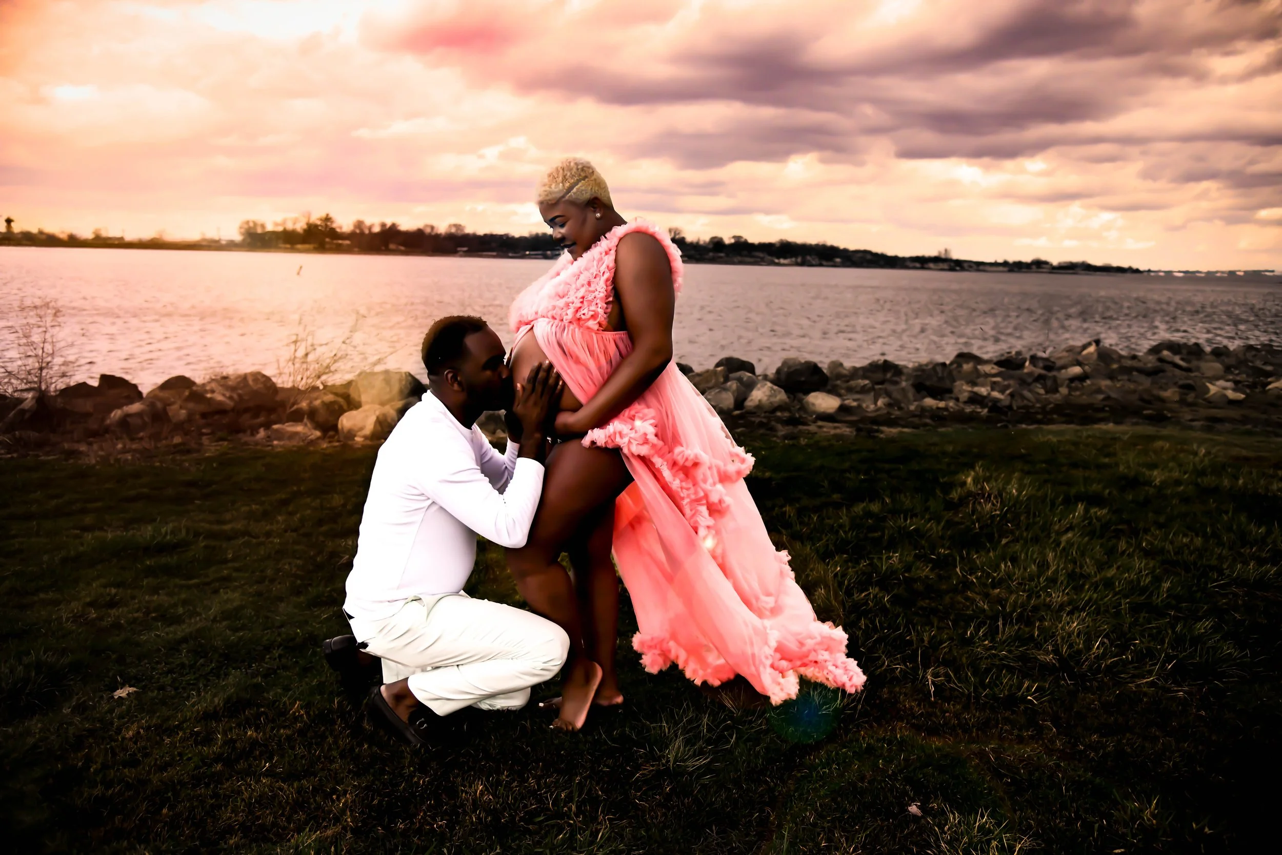A man kneeling and kissing a pregnant woman by a lake at sunset, with the woman in a flowing pink dress and the man in white.