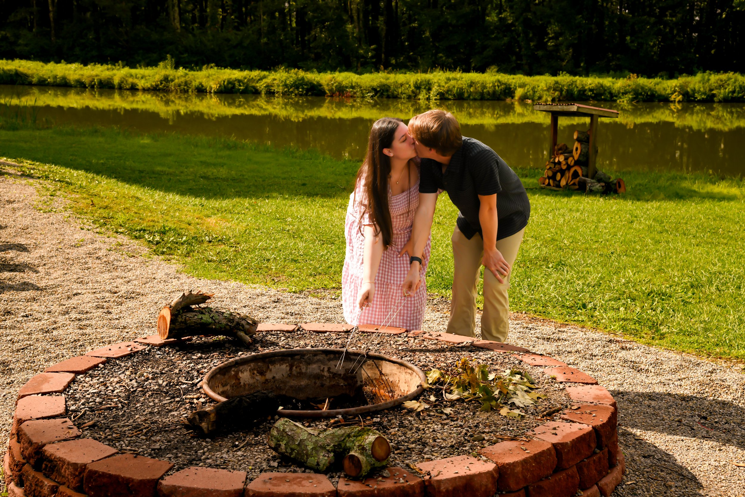 A young woman and a young man kissing near a campfire ring by a lake in a forested area during the day.