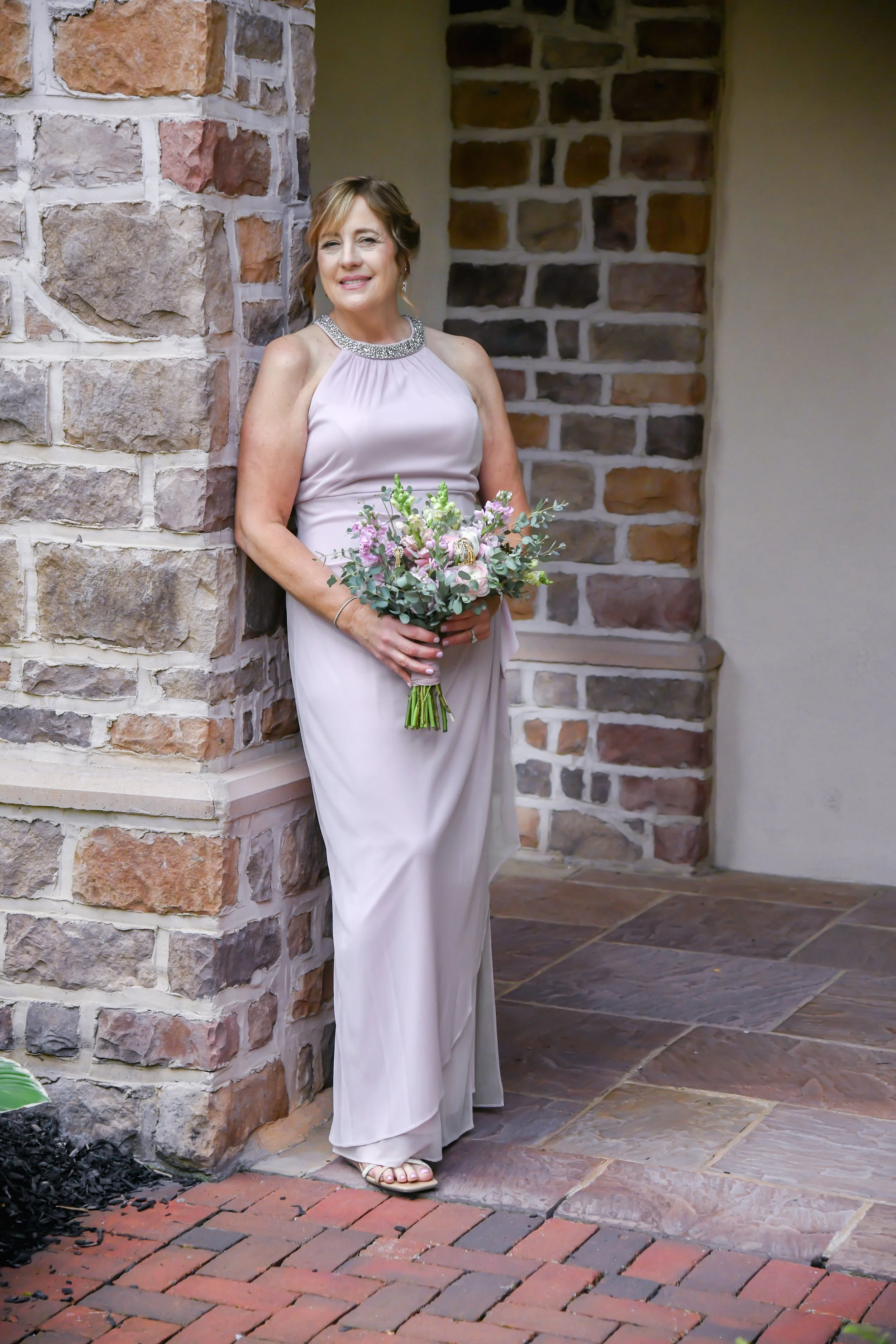 A woman in a light-colored formal dress holding a bouquet of flowers, standing against a brick wall.