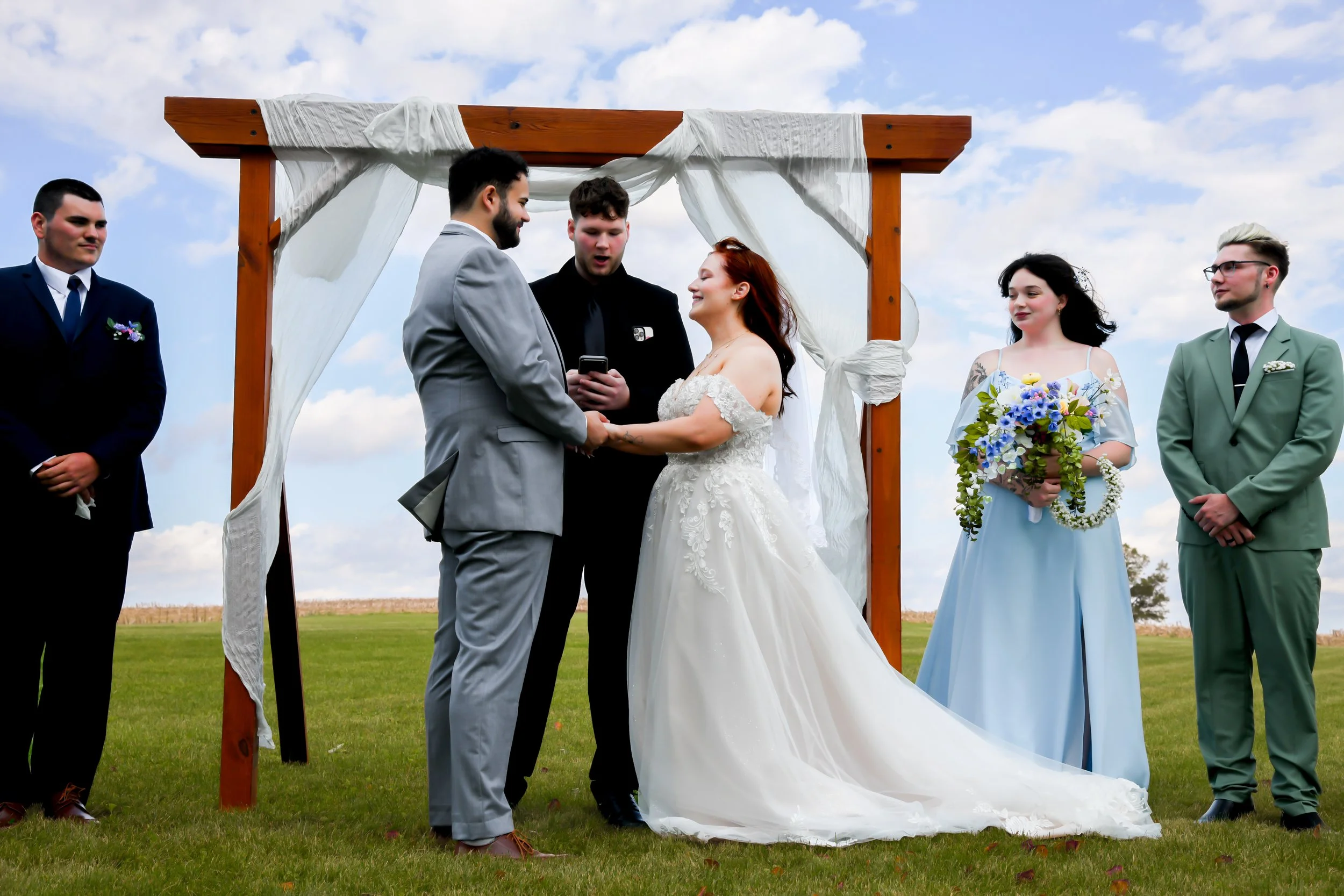 A wedding ceremony outdoors with seven people standing under a wooden arch with white drapes, a couple exchanging vows, and a man officiating.