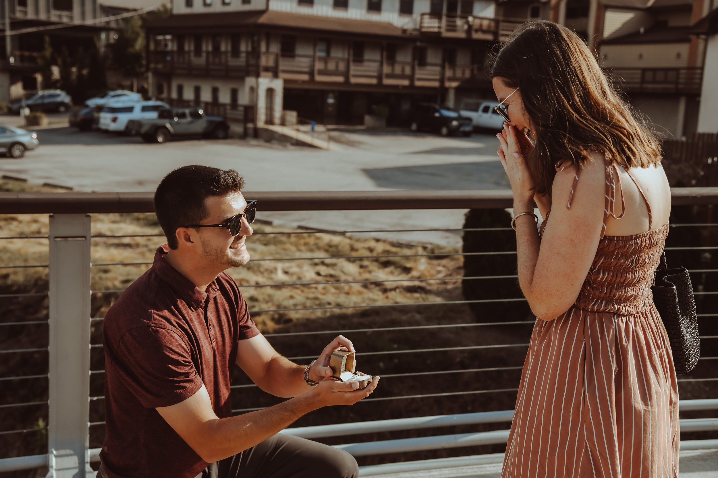 A man is proposing marriage to a woman on a bridge, holding an open ring box, while she reacts with surprise and her hands covering her mouth.