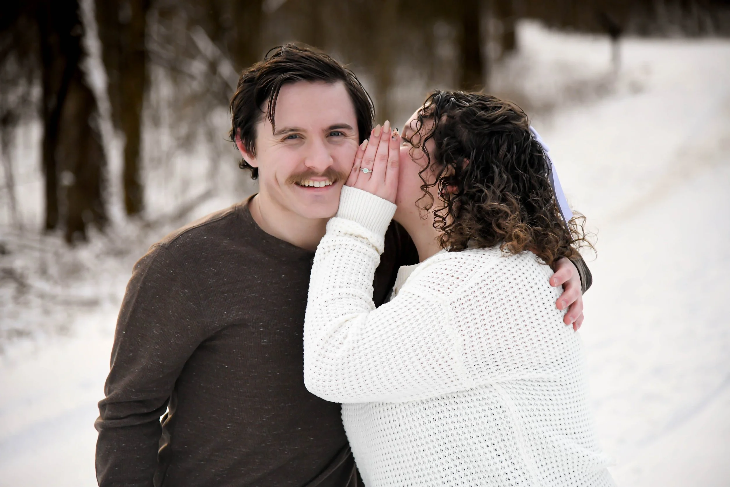 A couple standing outdoors in a snowy landscape, with the woman whispering into the man's ear and holding his face, both smiling.
