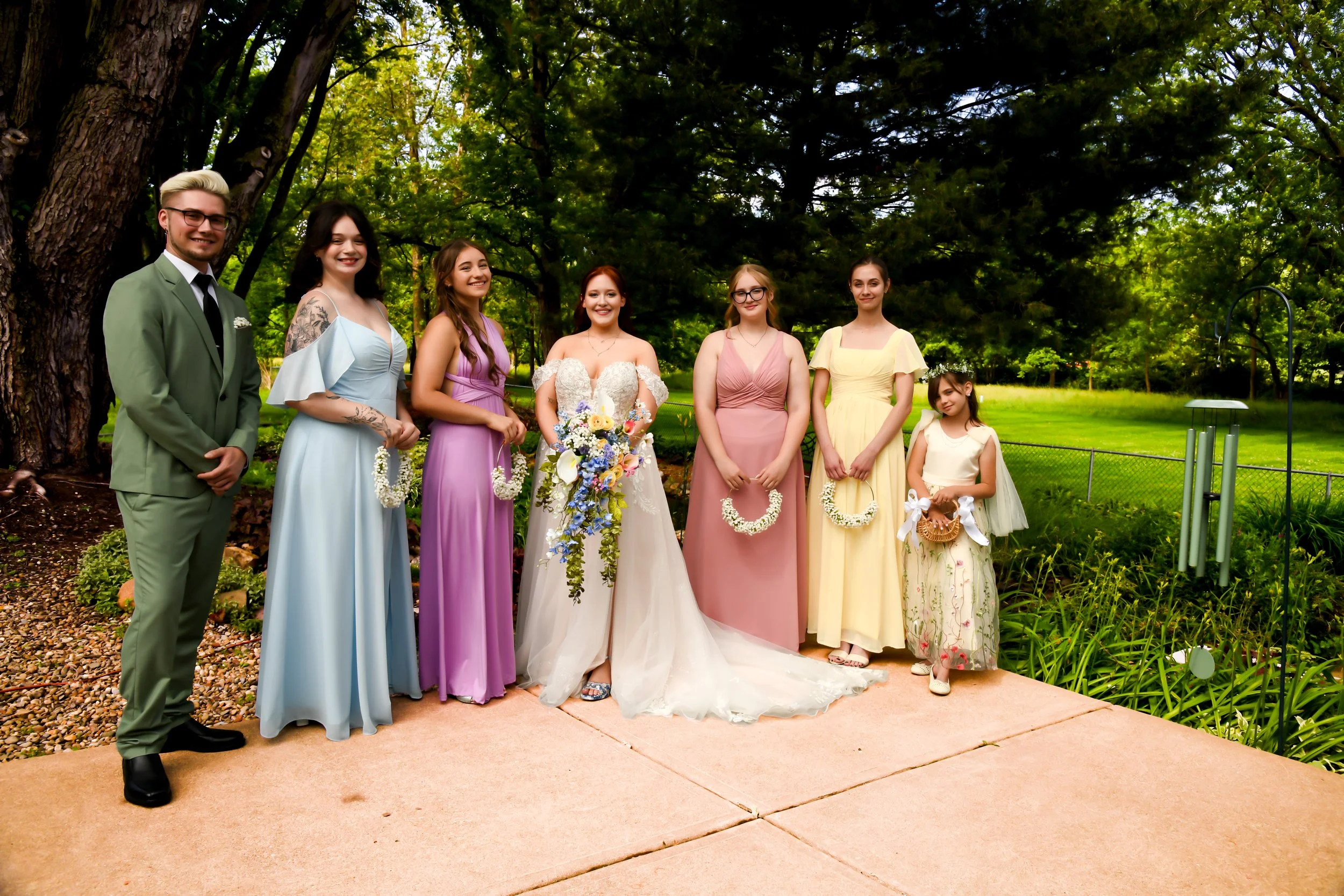A group of eight people, including adults and a child, dressed in formal and semi-formal attire, standing outdoors under large trees in a park-like setting with greenery and a cloudy sky.