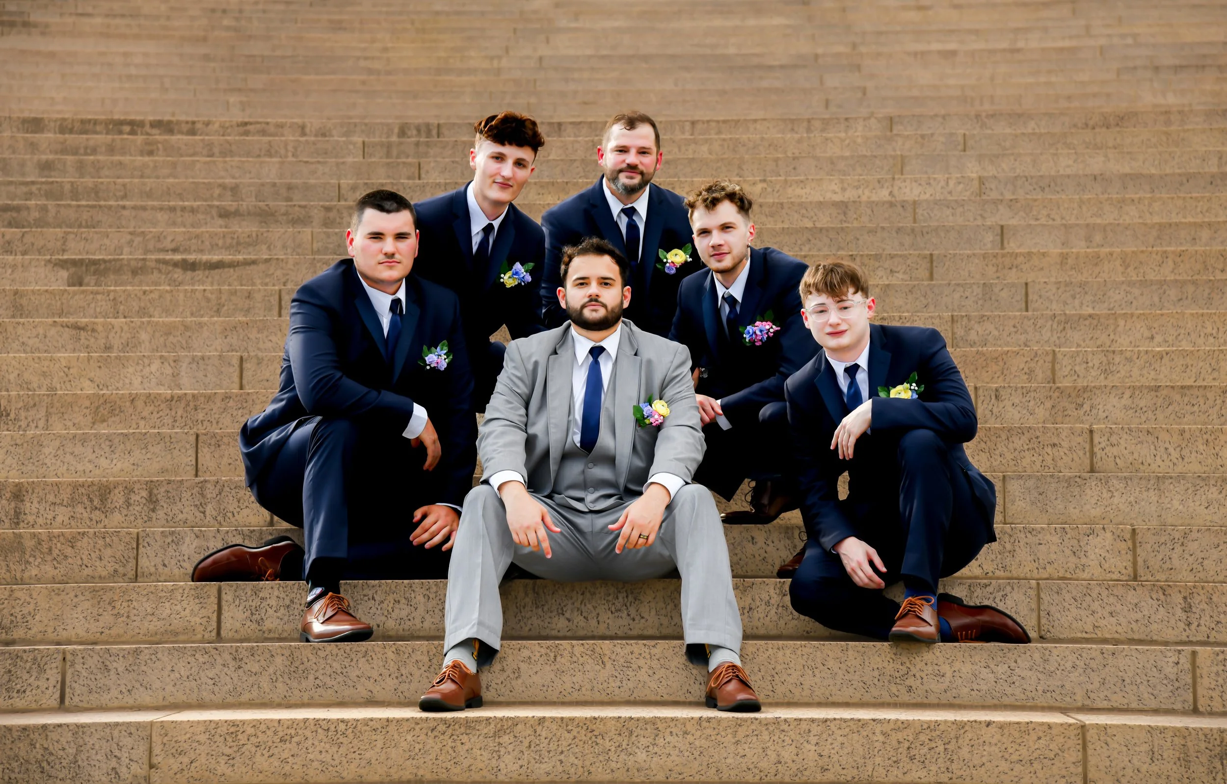 Group of seven men dressed in suits, posing on outdoor stairs, one sitting in front, others standing behind, all wearing boutonnières and formal shoes.