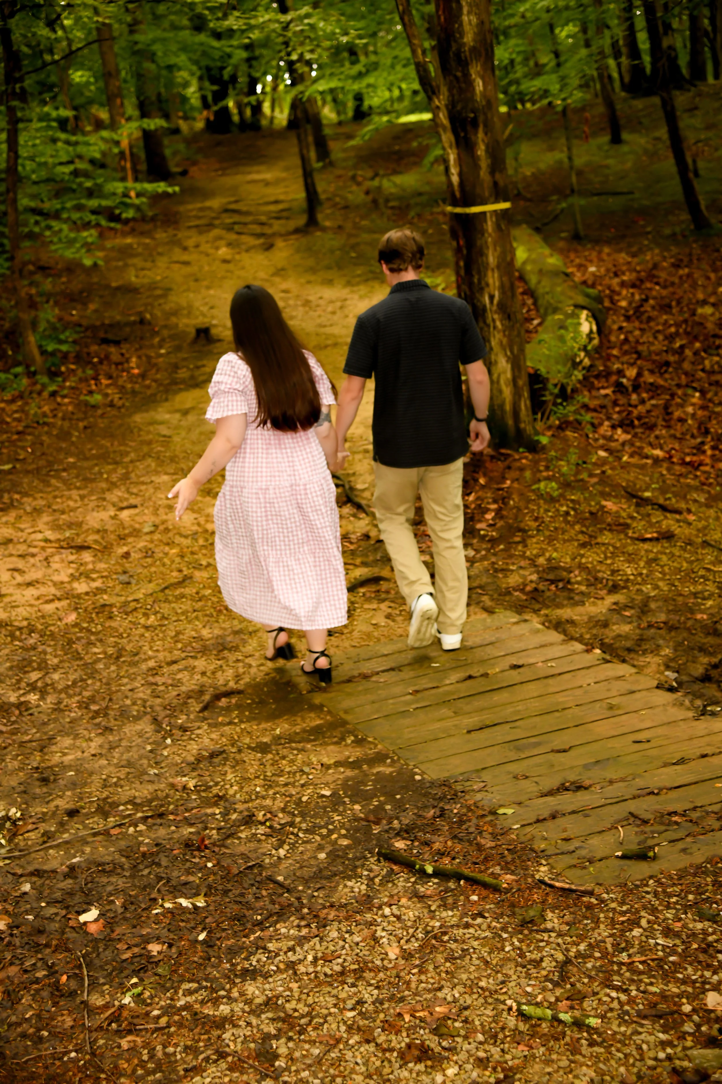 A couple walking hand in hand on a forest trail surrounded by trees with autumn leaves.