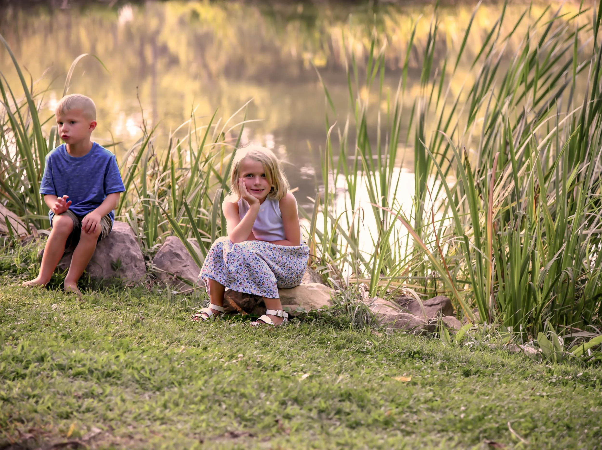 Two young children, a boy and a girl, sitting on rocks near a pond with tall grass and trees in the background during sunset.
