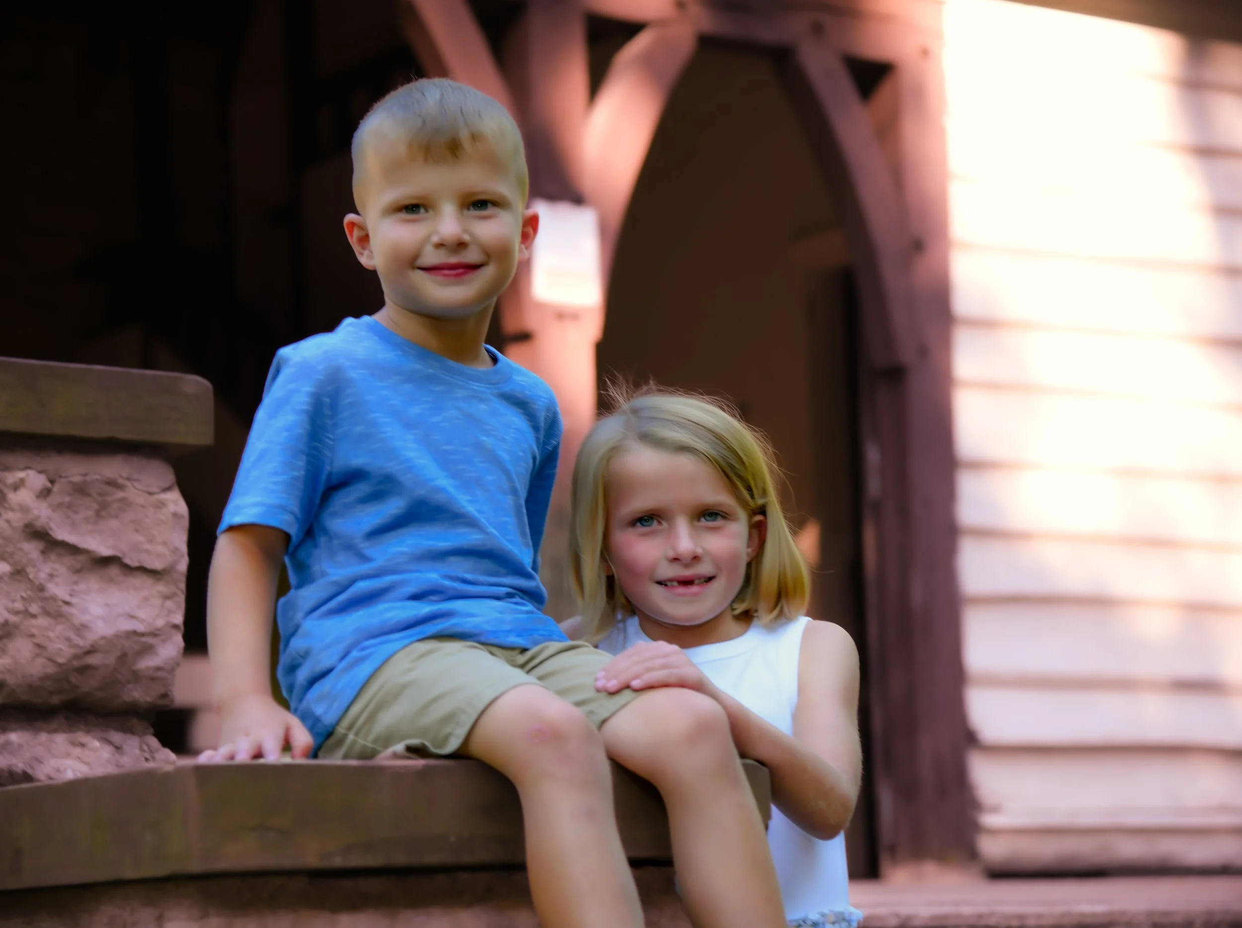 A young boy and girl sitting on a wooden ledge outdoors, smiling at the camera with a house or cabin in the background.