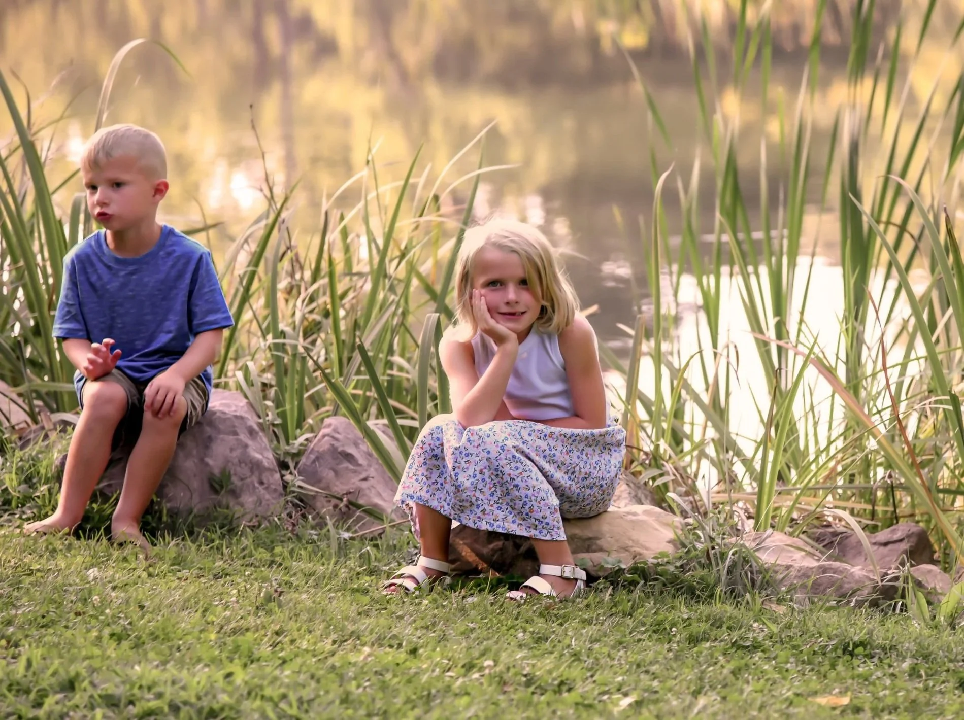 A young girl with long brown hair in a white floral dress is standing outdoors, holding onto a tree branch with one hand and smiling at the camera, surrounded by green trees and grass.