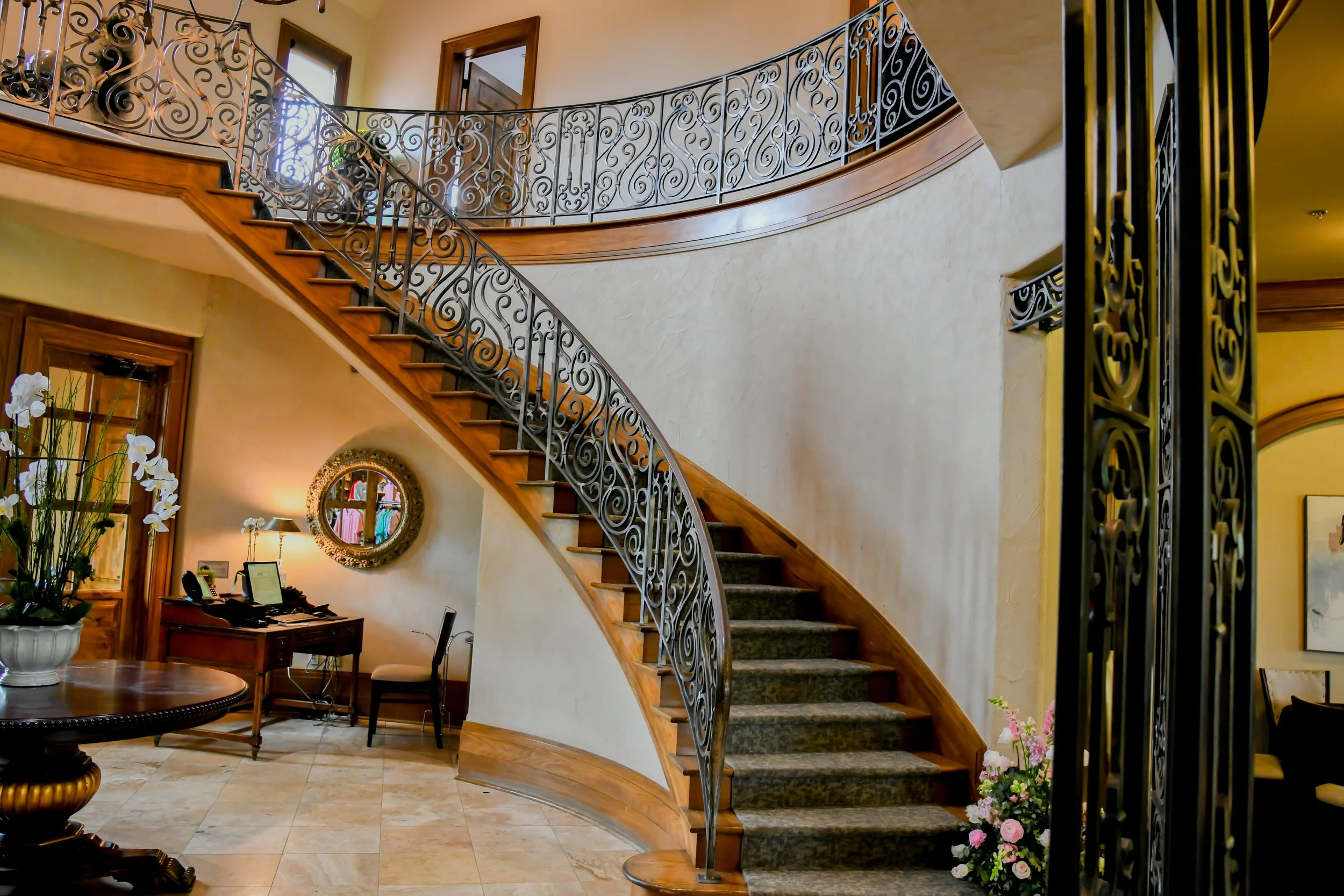 Interior of a luxurious home with a curved staircase featuring ornate black wrought iron railing, wood steps with a carpet runner, and a foyer decorated with flowers and furniture.