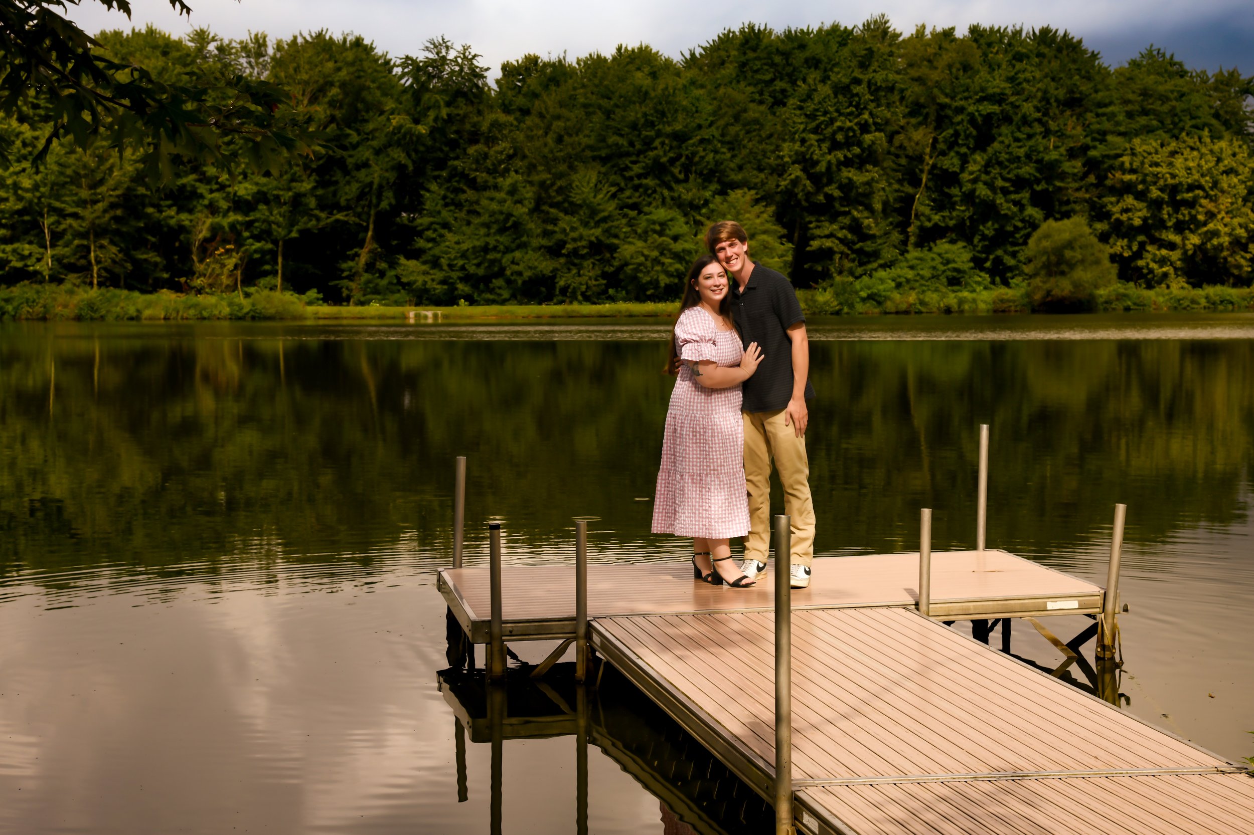 A smiling young couple standing as friends on a wooden dock by a lake with calm water, surrounded by green trees and a cloudy sky.