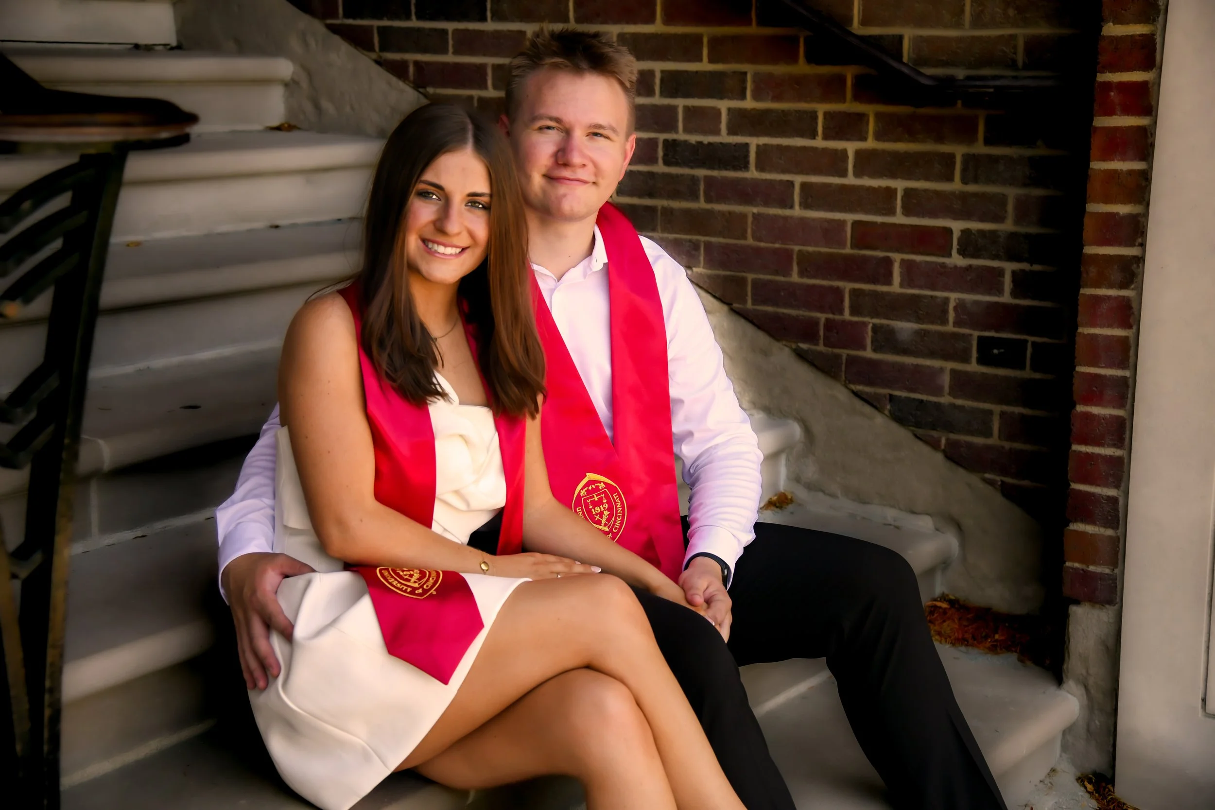 Two young graduates, a woman and a man, sitting on concrete steps outside, smiling, wearing red graduation sashes.