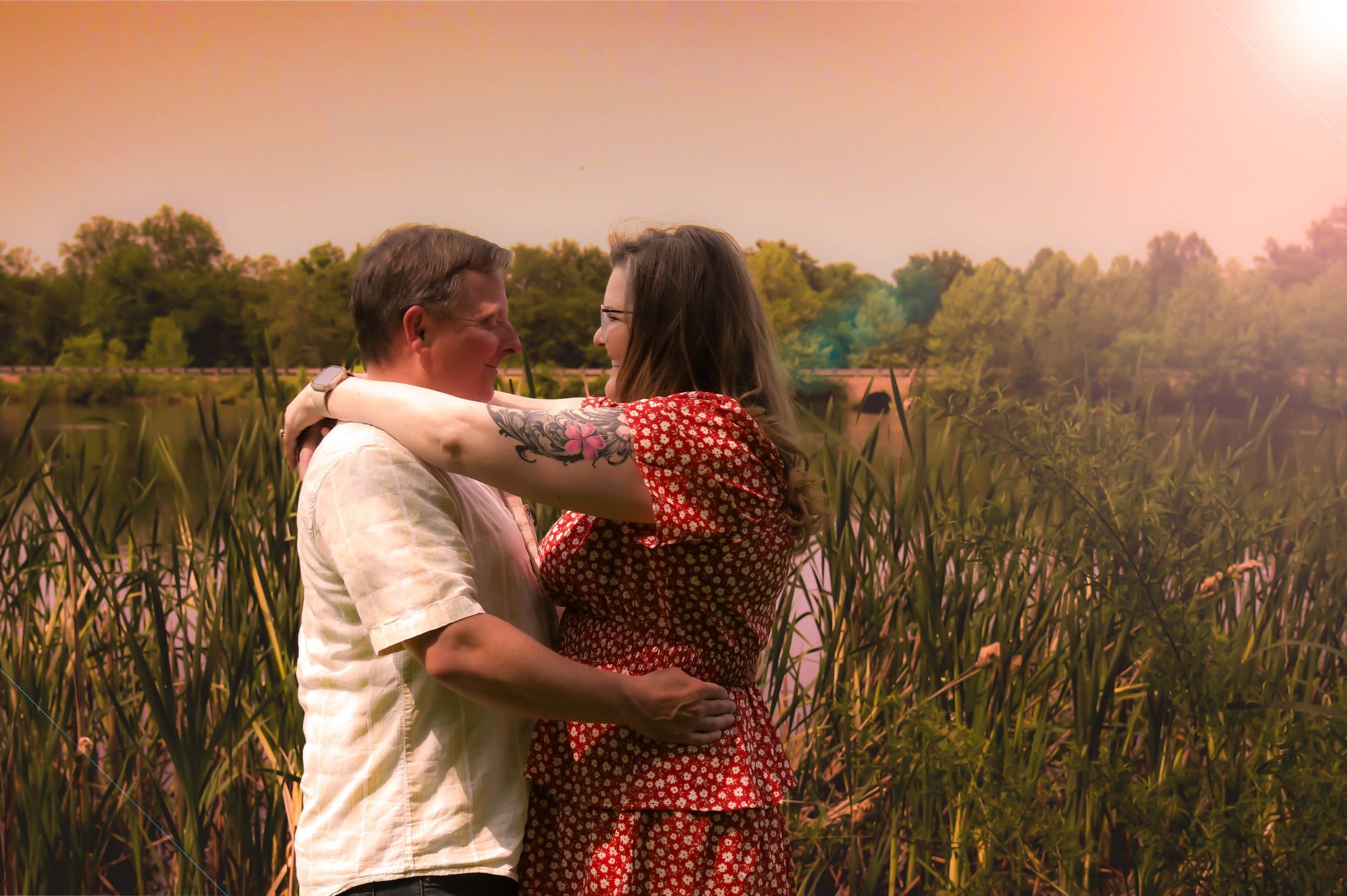 A couple embracing and smiling at each other outdoors near a body of water with trees in the background.