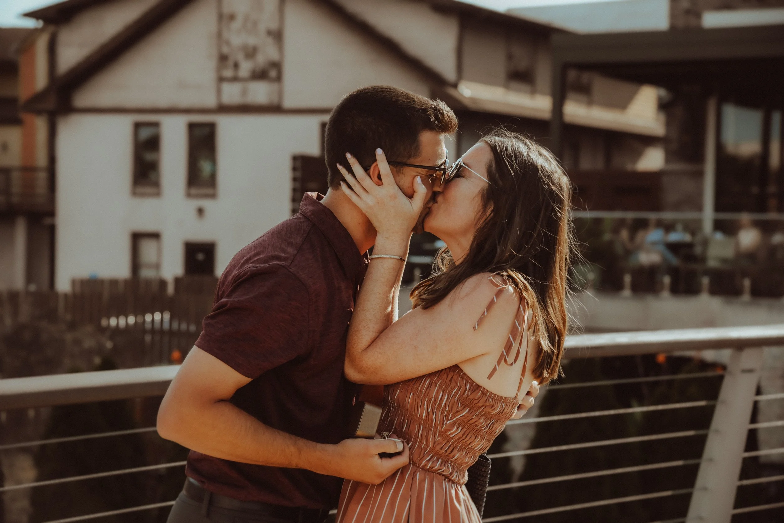 A couple sharing a kiss outside on a sunny day, with a railing and house in the background.