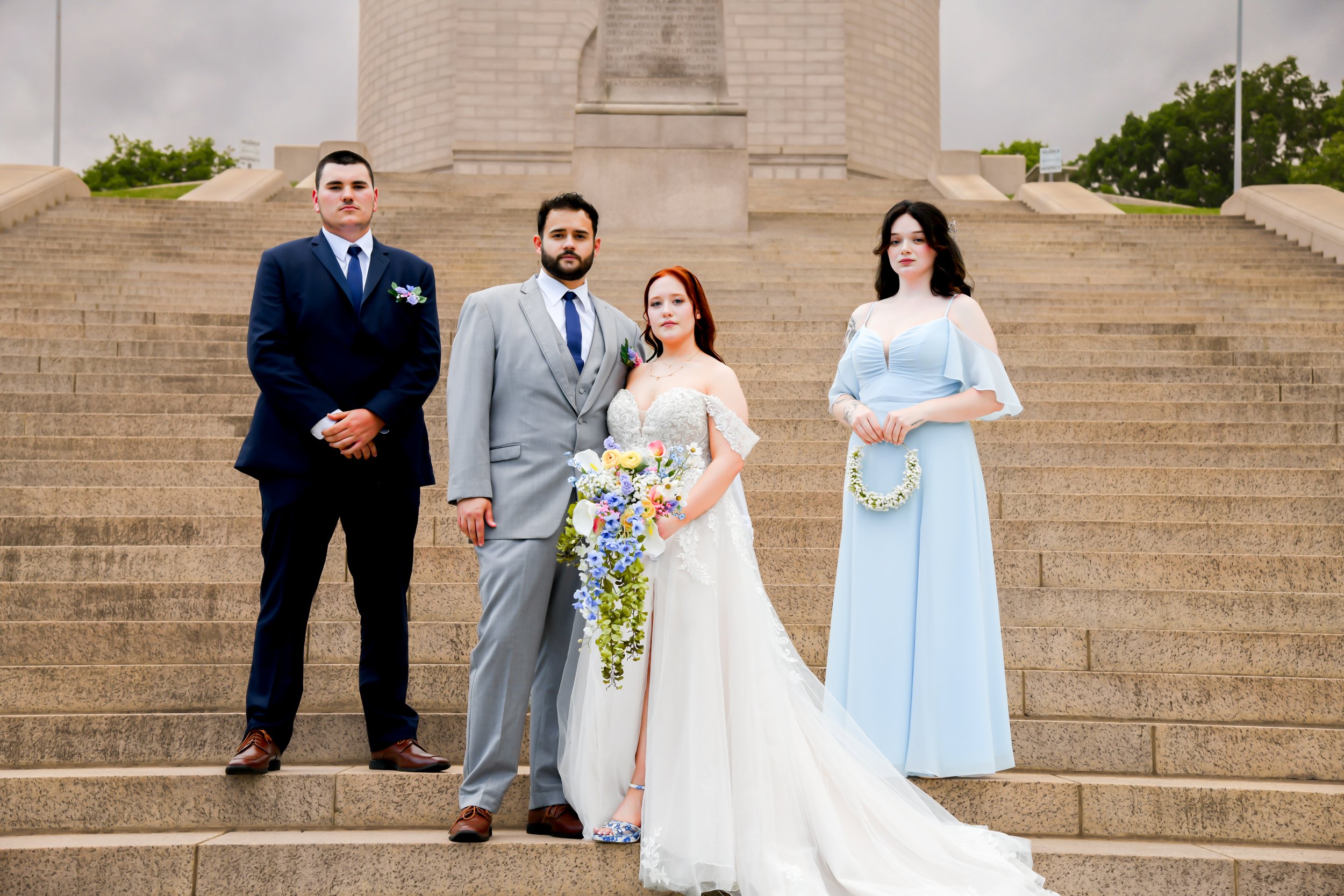 Group of four people in formal attire standing on steps in front of a large stone monument, the setting appears to be a city or park, overcast sky.