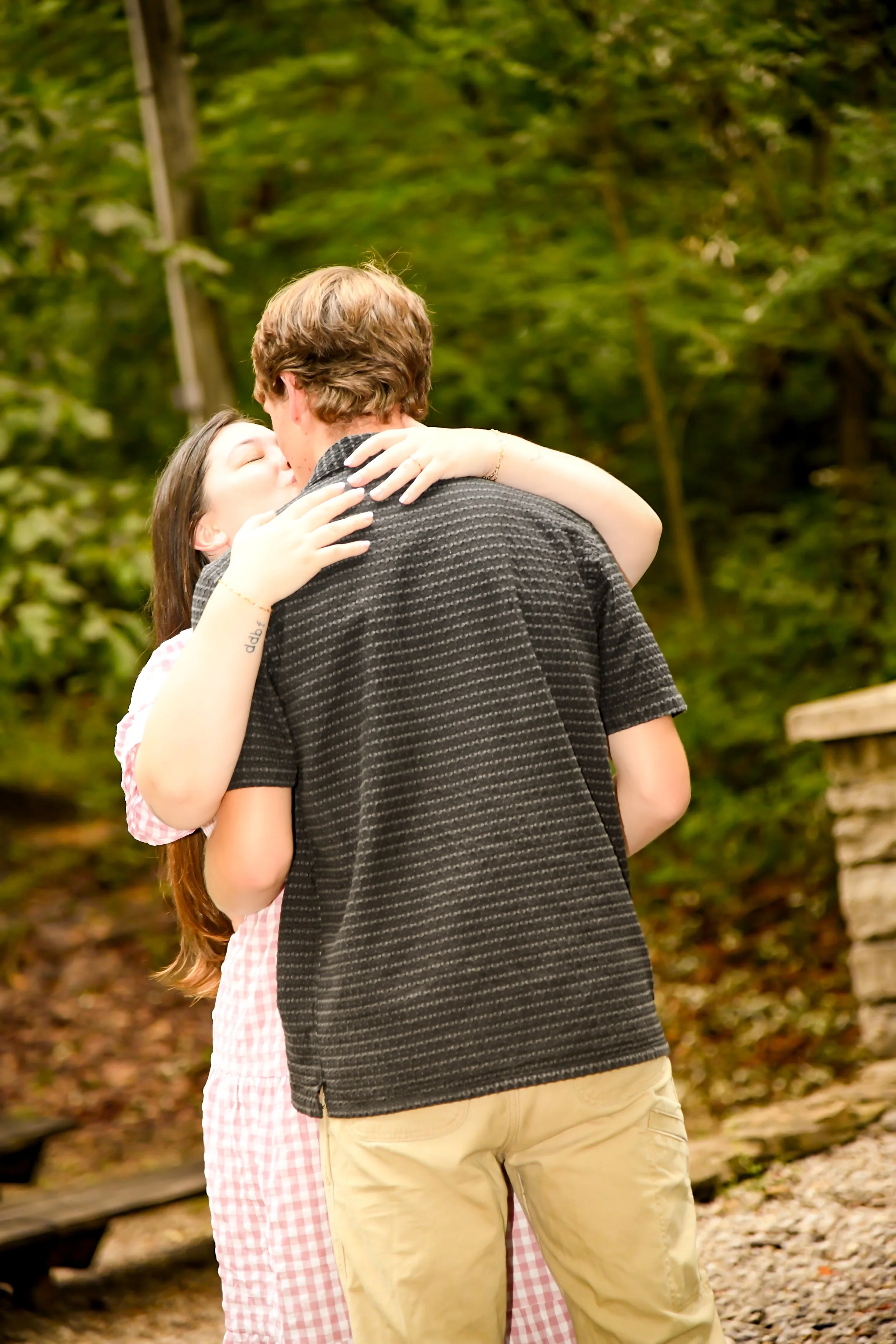 A young woman and a young man share a kiss outdoors in a wooded area.