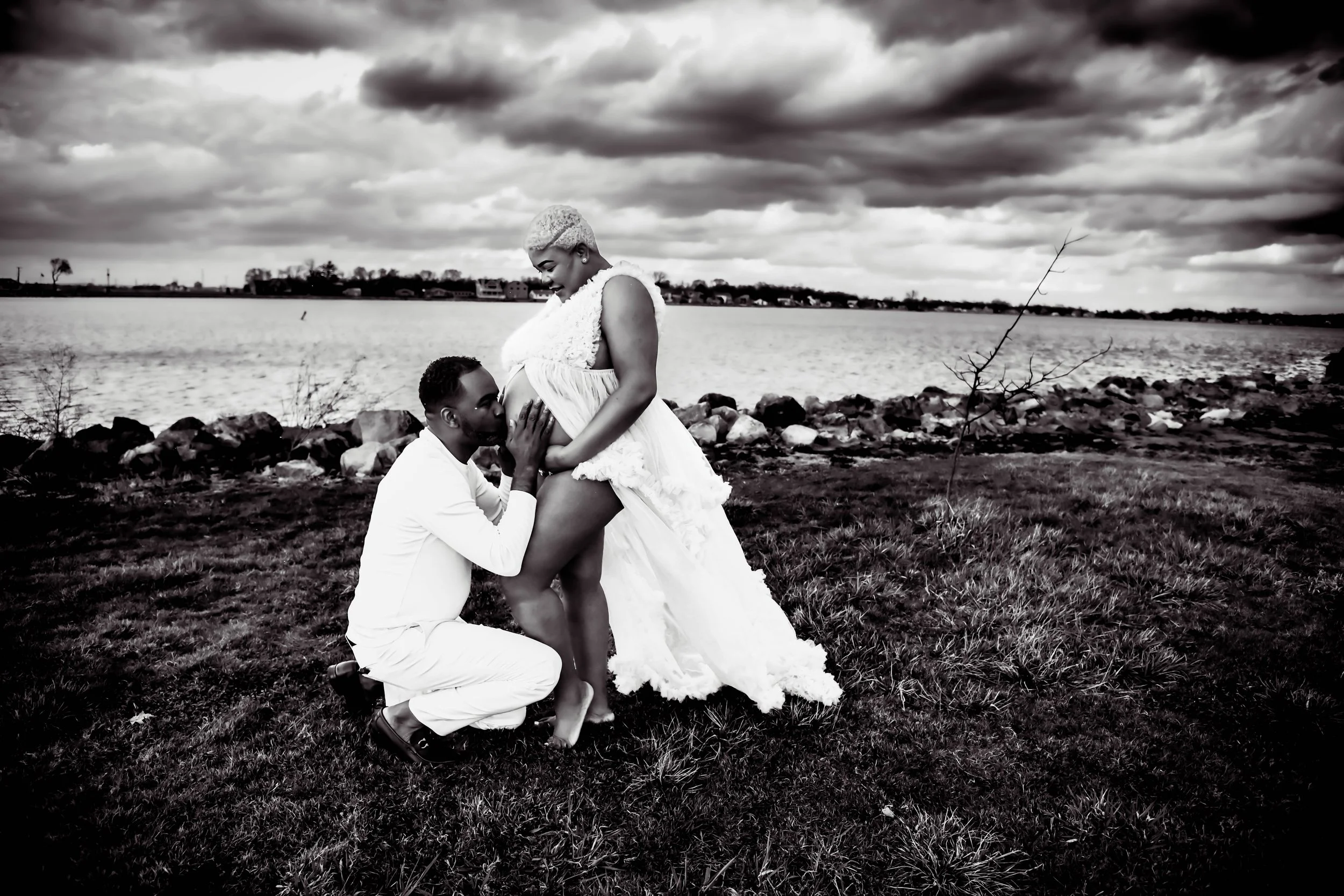 A black and white photo of a couple by a lake. The man is kneeling, kissing the pregnant woman's belly. The woman is standing, wearing a flowing dress and smiling, with clouds in the sky and rocks near the shore.